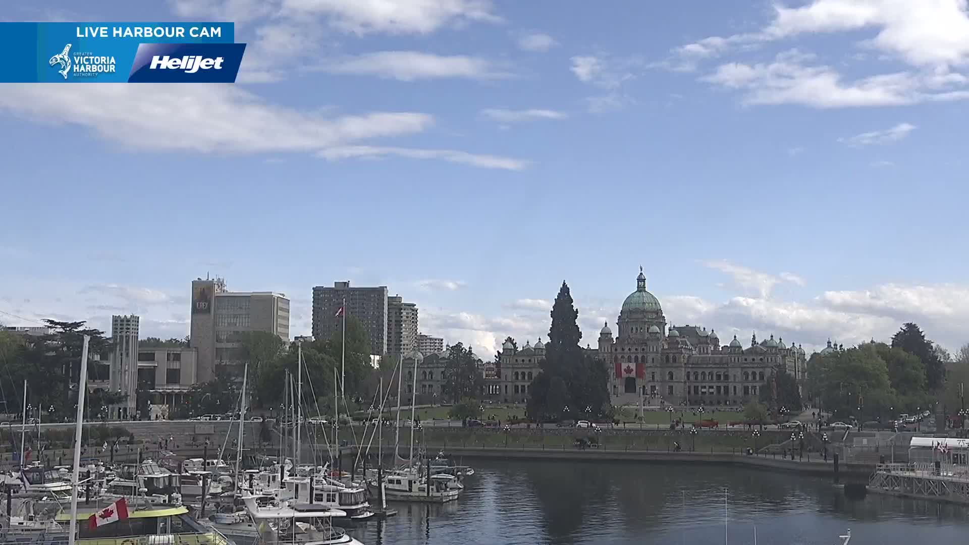 A harbor filled with sailboats is in front of a city skyline featuring a large, domed building under a mostly sunny sky with scattered clouds.