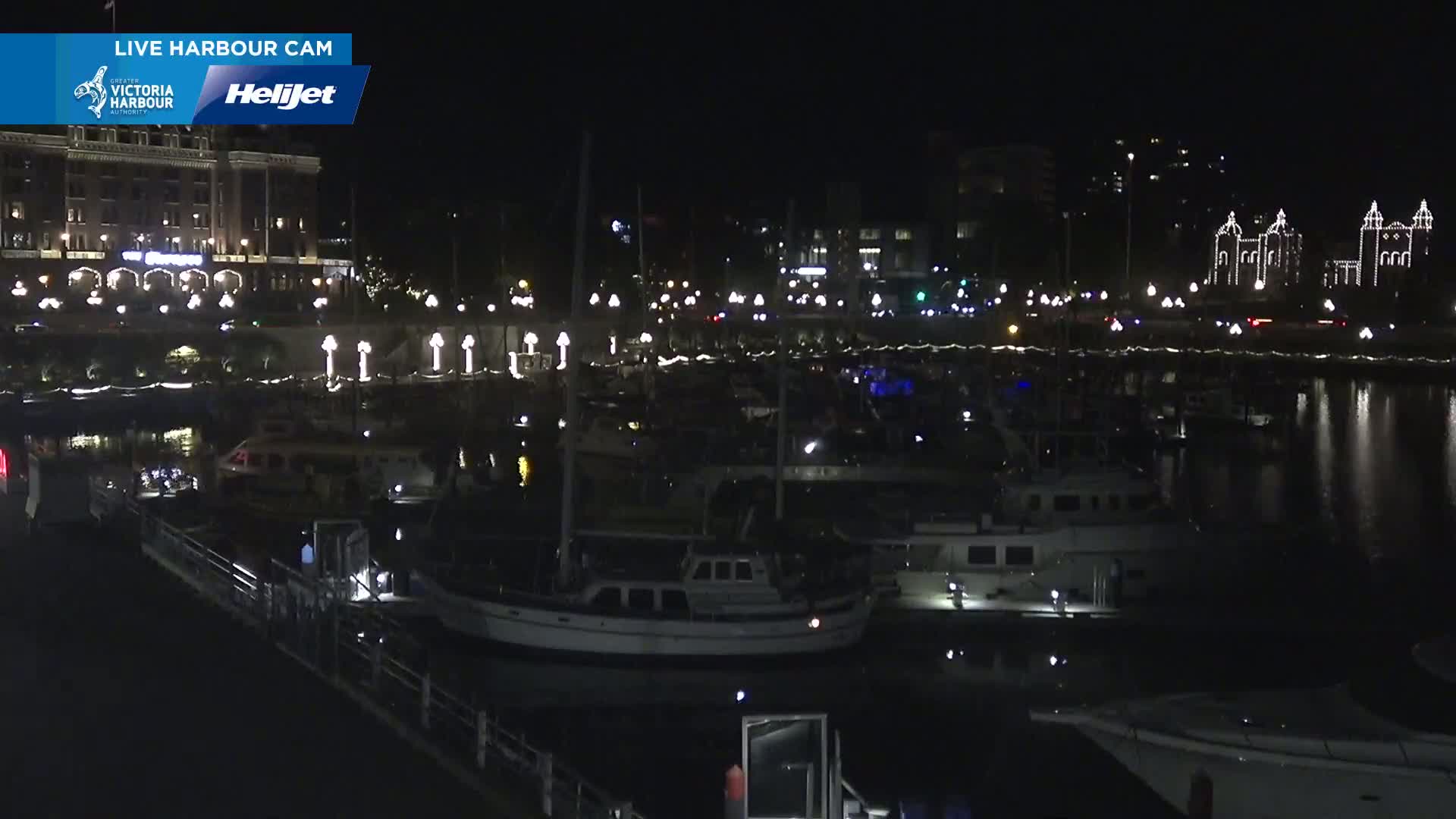 A nighttime view of a harbor filled with boats moored along a waterfront, featuring a large building illuminated with lights in the background under a clear sky.