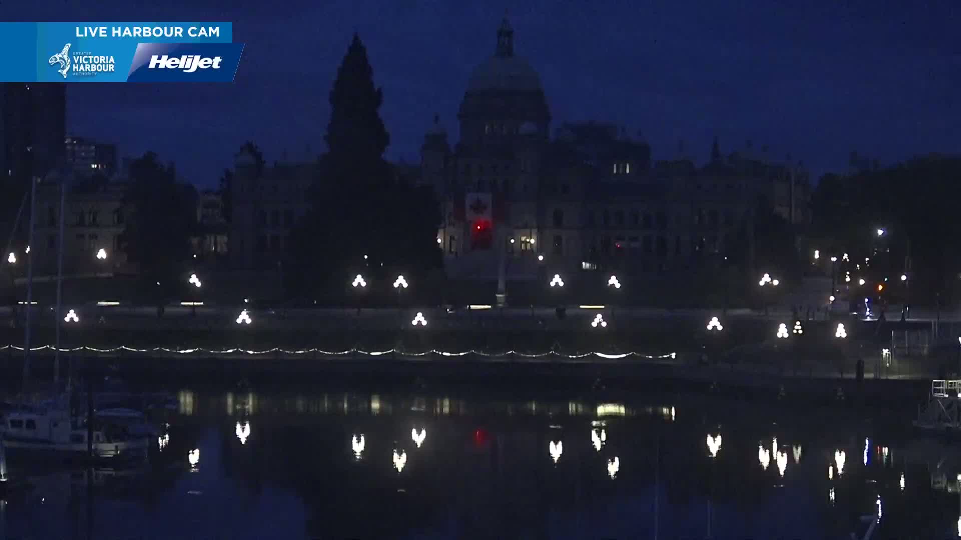 At night, a large, dark building with a dome is illuminated by several lights along a waterfront, reflecting in calm water where boats are moored.