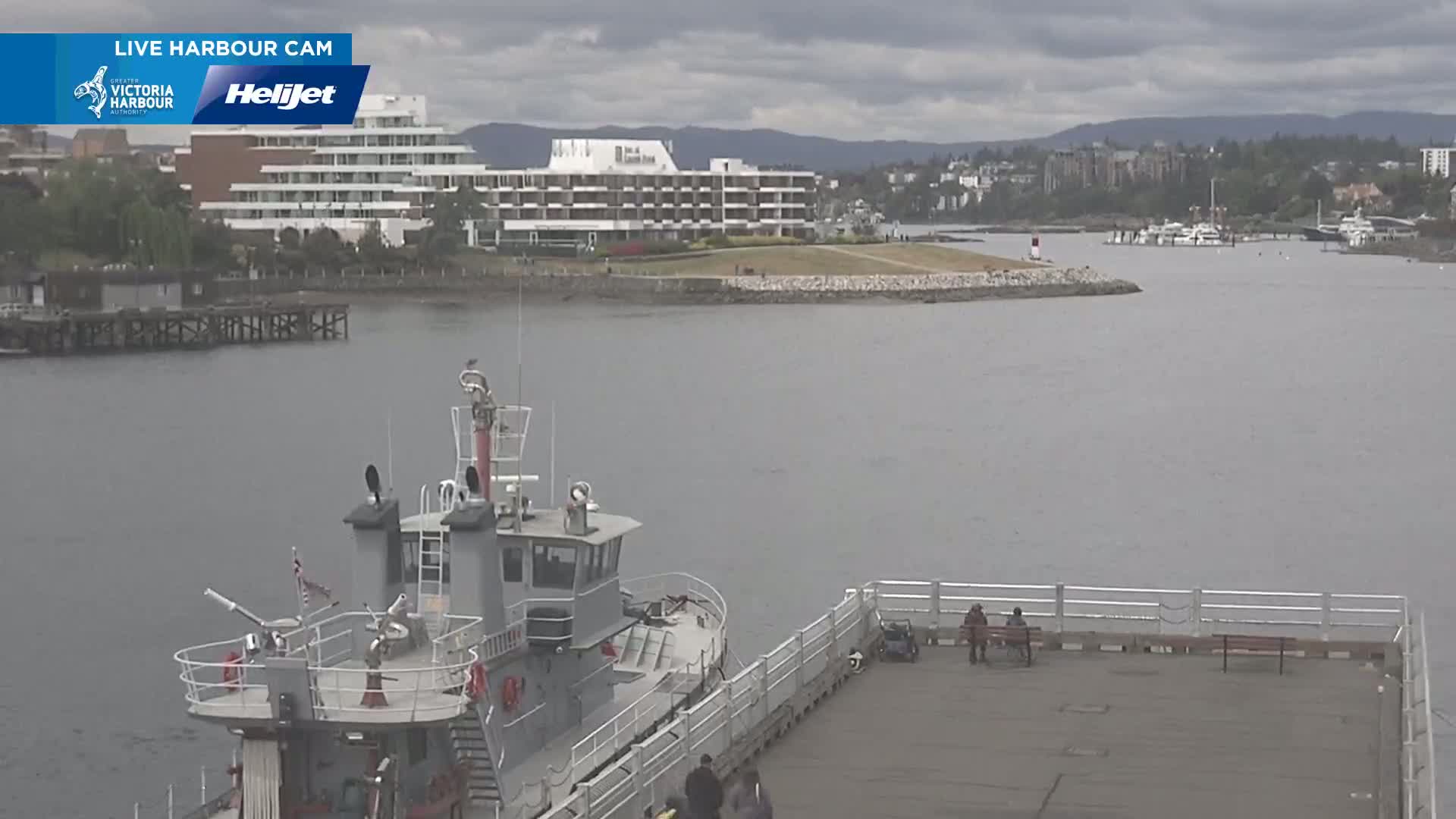 A grey fireboat is moored at a pier next to calm grey water, with a large white hotel and other buildings visible in the background under an overcast sky.