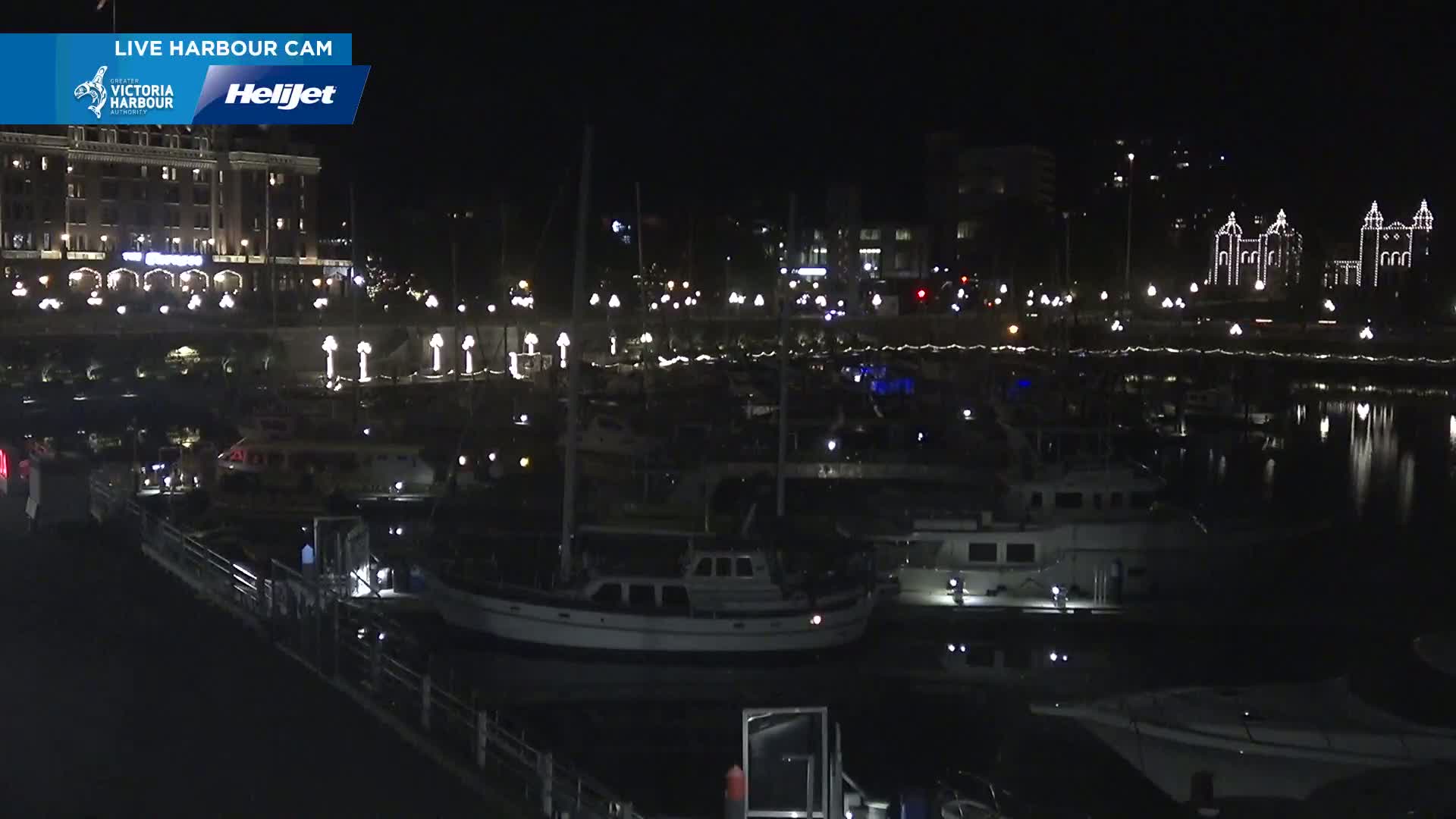 At night, a harbor filled with boats is illuminated by lights on buildings and along the docks.
