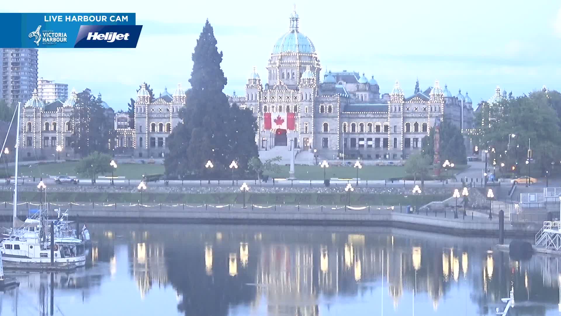 A large, illuminated stone building with a dome, flying a Canadian flag, sits beside a calm body of water, reflecting the building and lights at twilight.