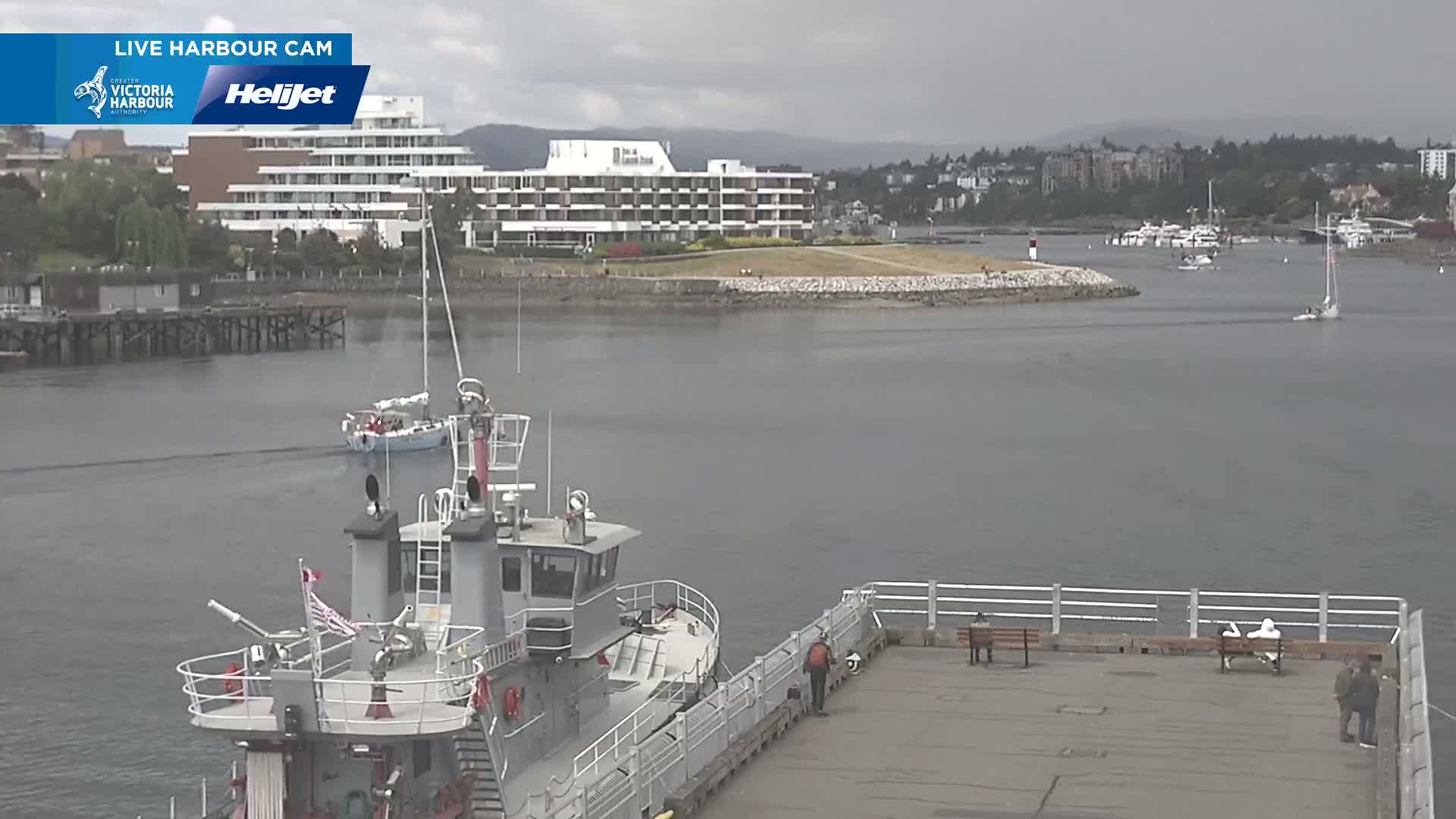 A gray harbor patrol boat is docked at a pier, overlooking calm gray water with several sailboats and a large hotel in the background under an overcast sky.