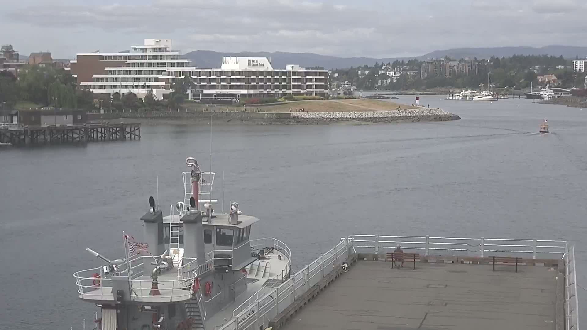 A gray fireboat is docked at a pier overlooking a calm body of water with a large hotel and a small town in the background under an overcast sky.