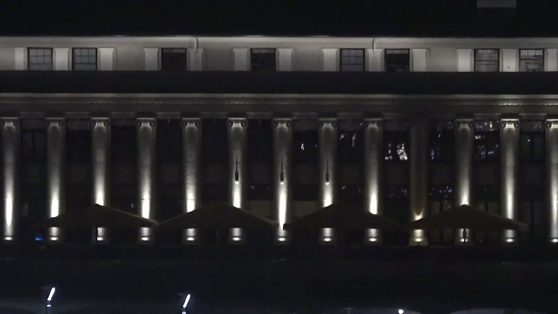 A large, classical-style building with columns is illuminated at night, and there are several small, covered outdoor seating areas in front of it.