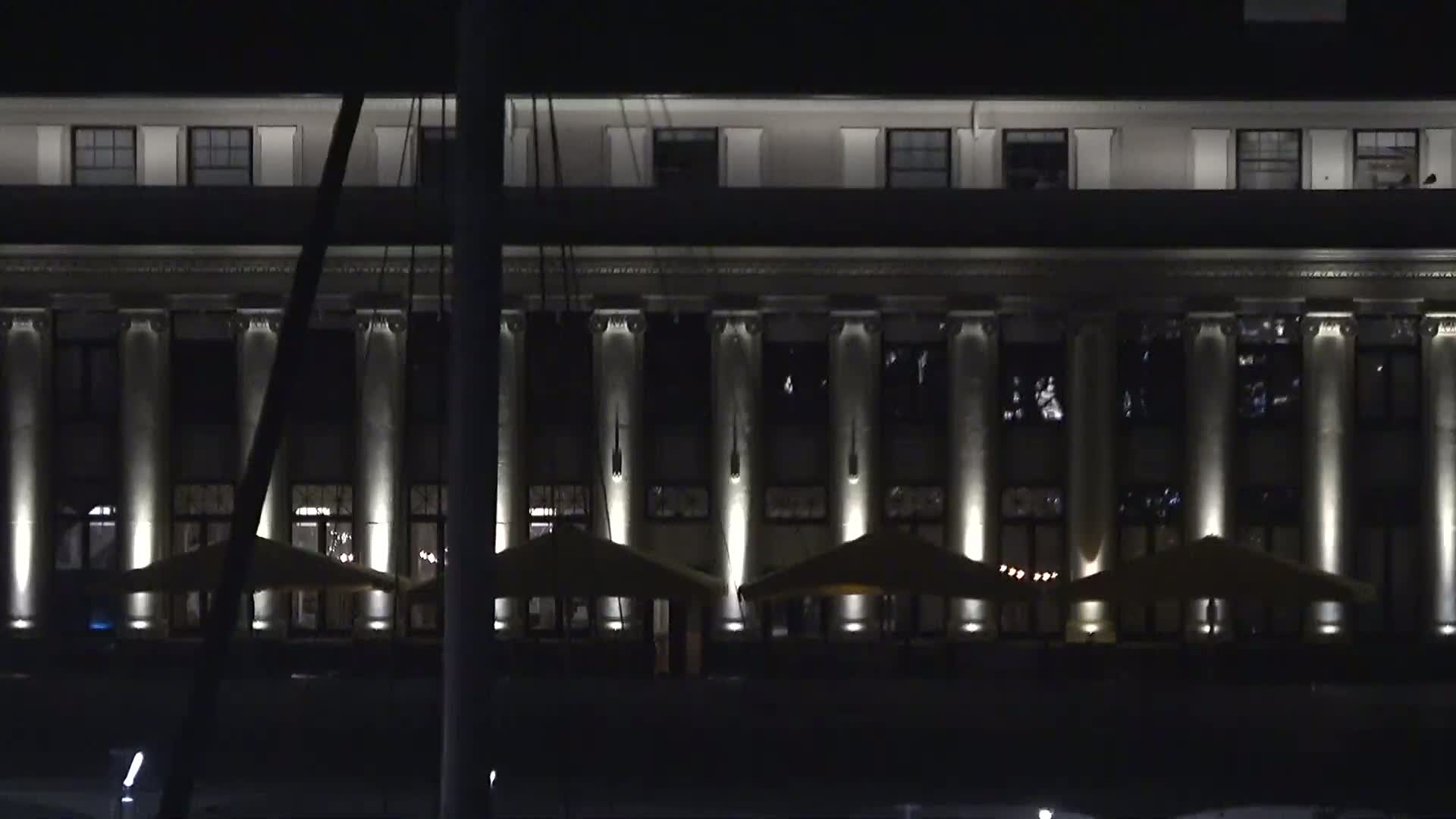 At night, a large, classically-styled building with columns is illuminated, and several outdoor patio umbrellas are set up in front.