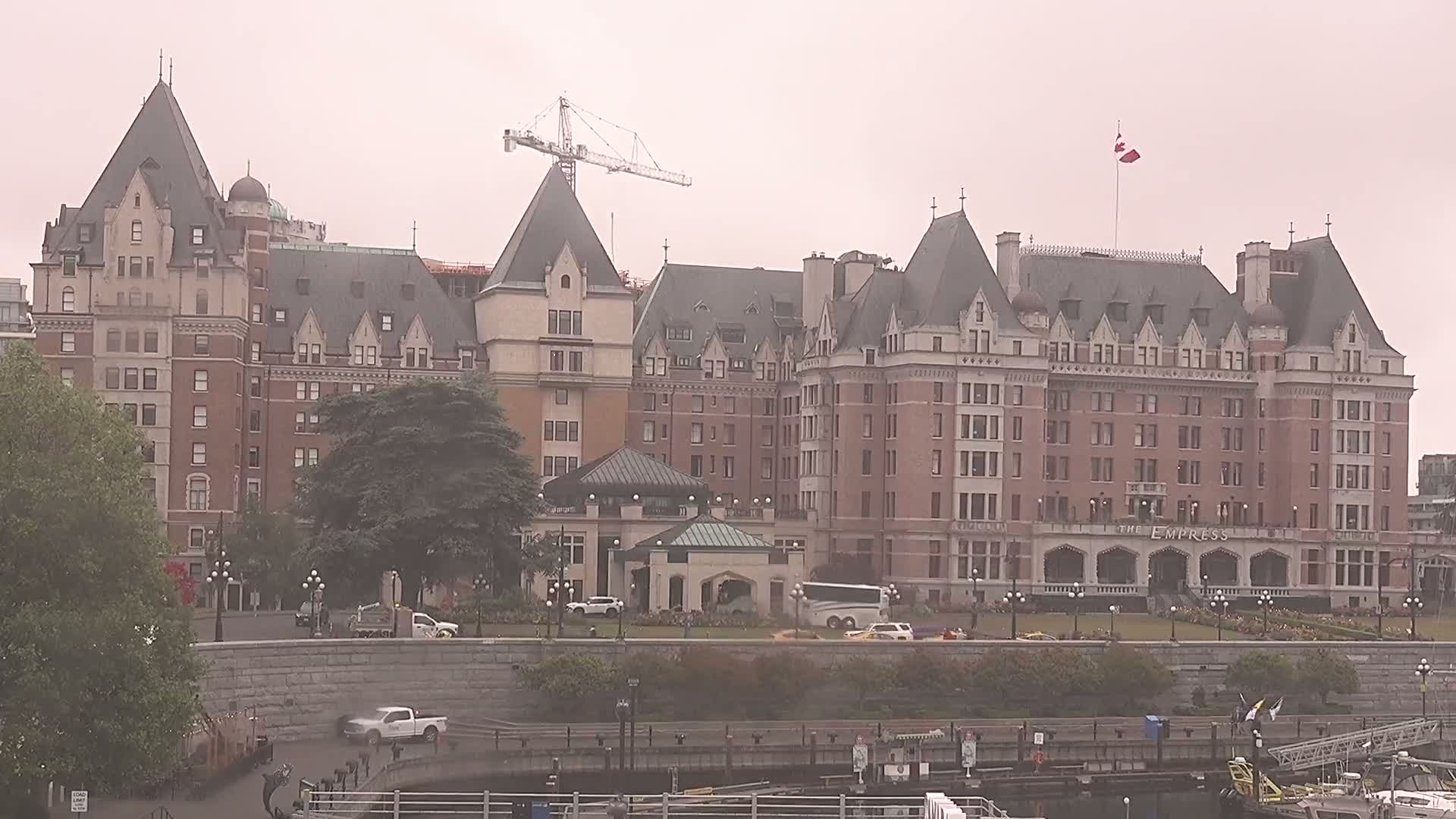 A large, brick chateau-style hotel with a Canadian flag flying atop, partially obscured by fog and trees, is seen from across a waterway, with a construction crane visible in the background.