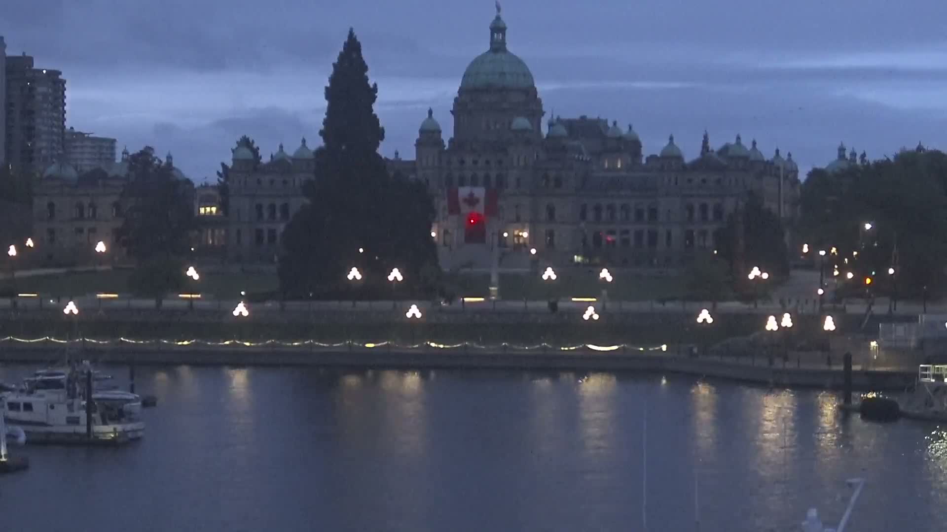 A large, stone building with a dome, illuminated at night, sits on the waterfront, with boats moored in the calm water under a dark, dusky sky.