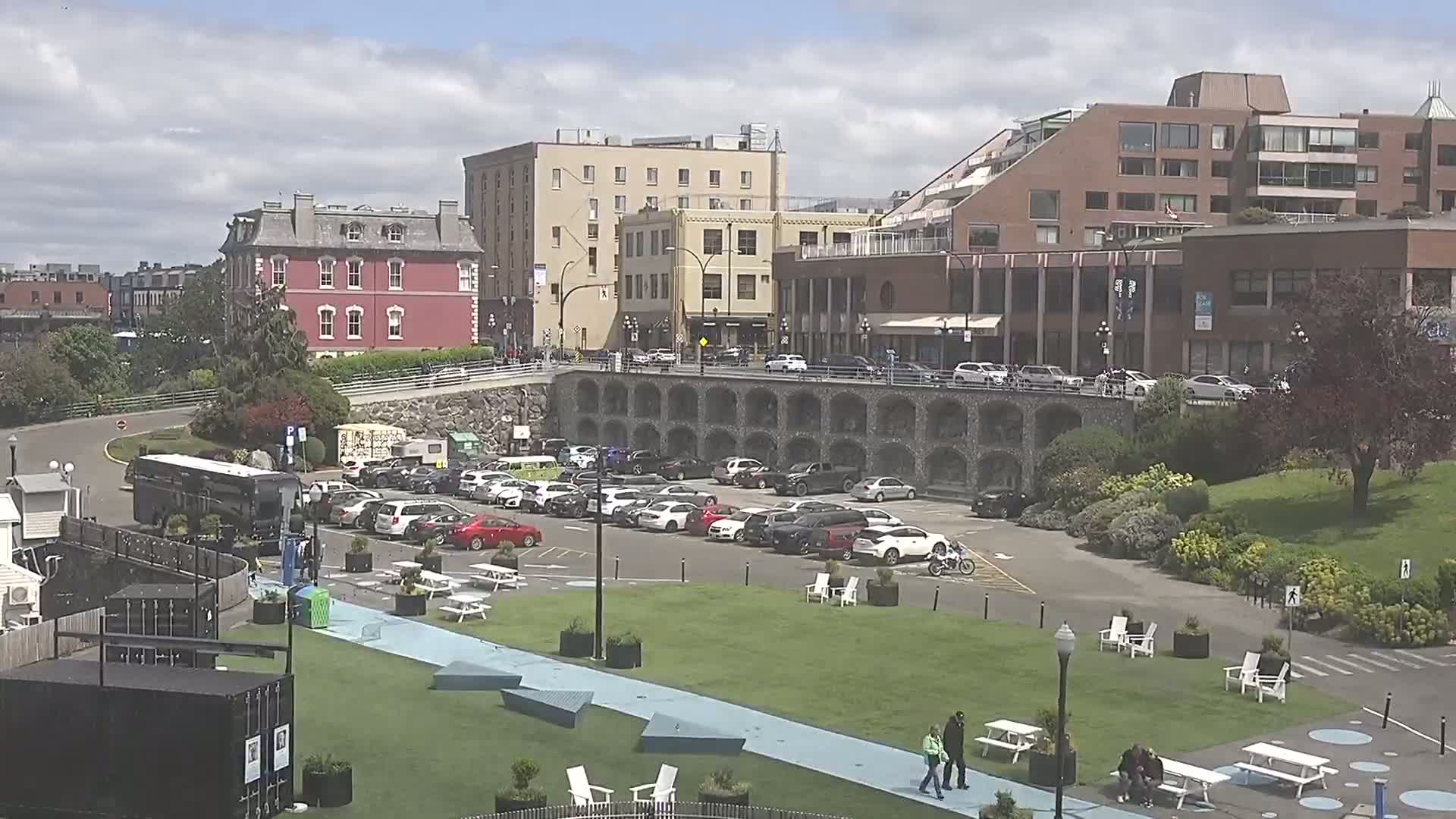 A partly cloudy day shows a city park with a paved area, a grassy lawn, picnic tables, and white chairs, next to a parking lot and several buildings.