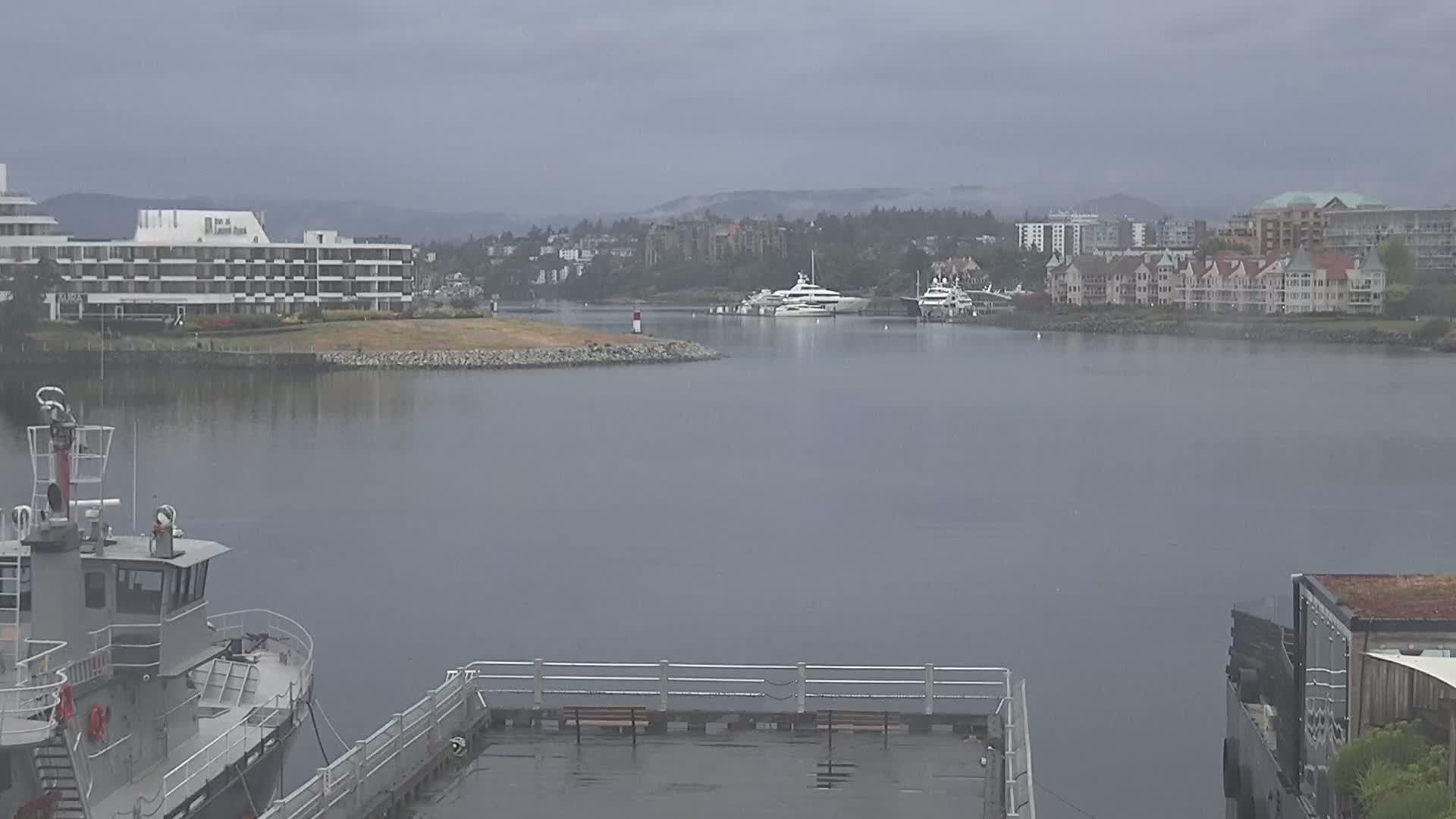 A calm harbor scene on an overcast day shows several yachts moored near a waterfront lined with buildings, viewed from a partially visible boat or dock.
