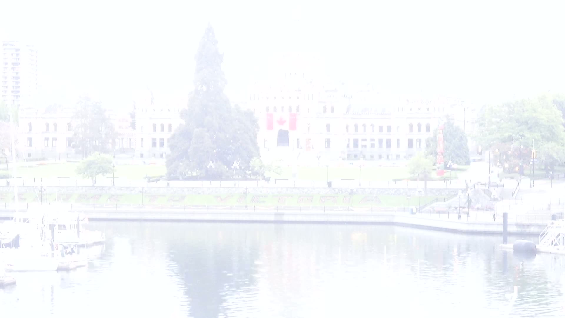 A hazy view shows a waterfront scene with boats docked, a grassy area, and a large stone building with Canadian flags displayed prominently in front.