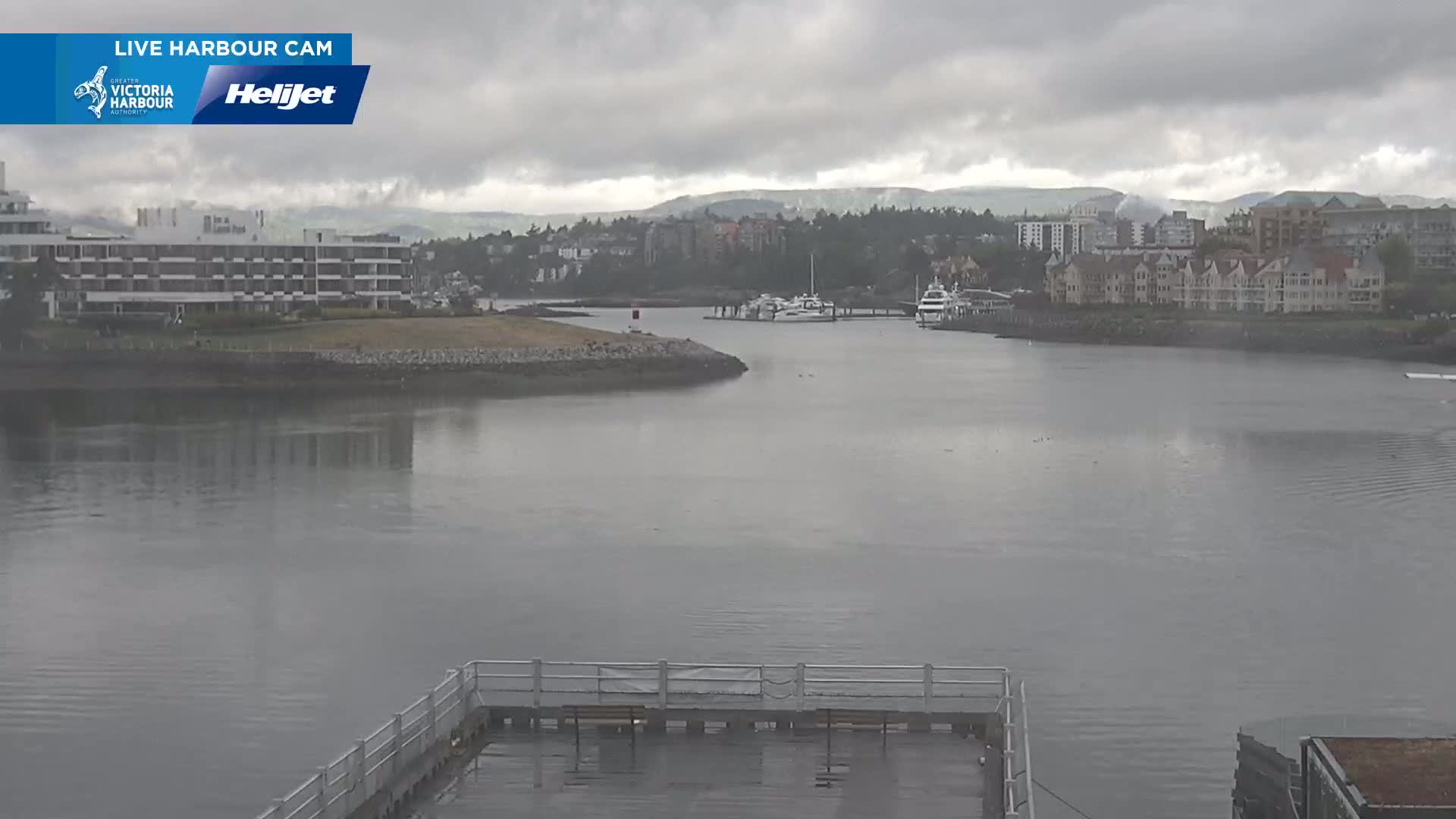 A harbor scene on an overcast day shows calm water, several boats docked, and buildings along the shoreline.