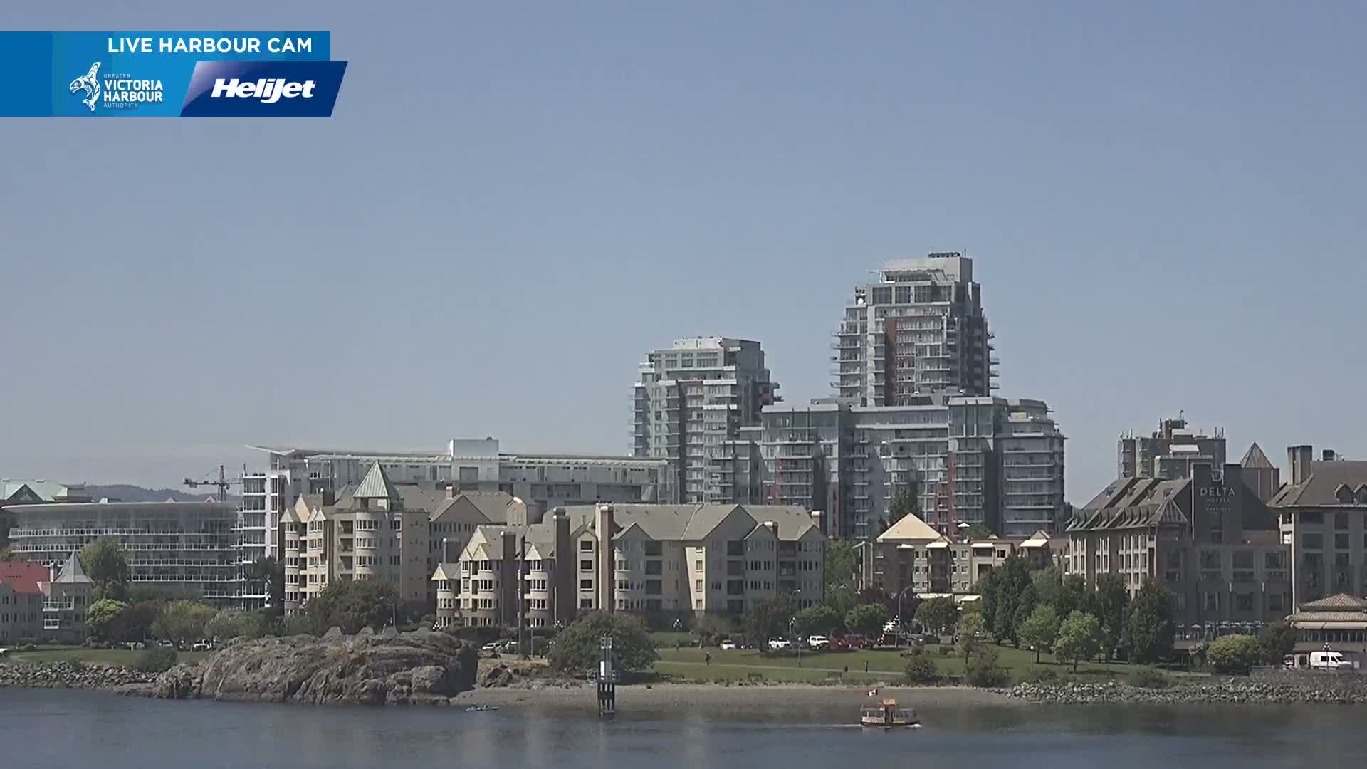 A waterfront scene on a clear, sunny day shows several multi-story buildings, including apartment complexes and hotels, situated along a shoreline with a rocky beach and calm water.