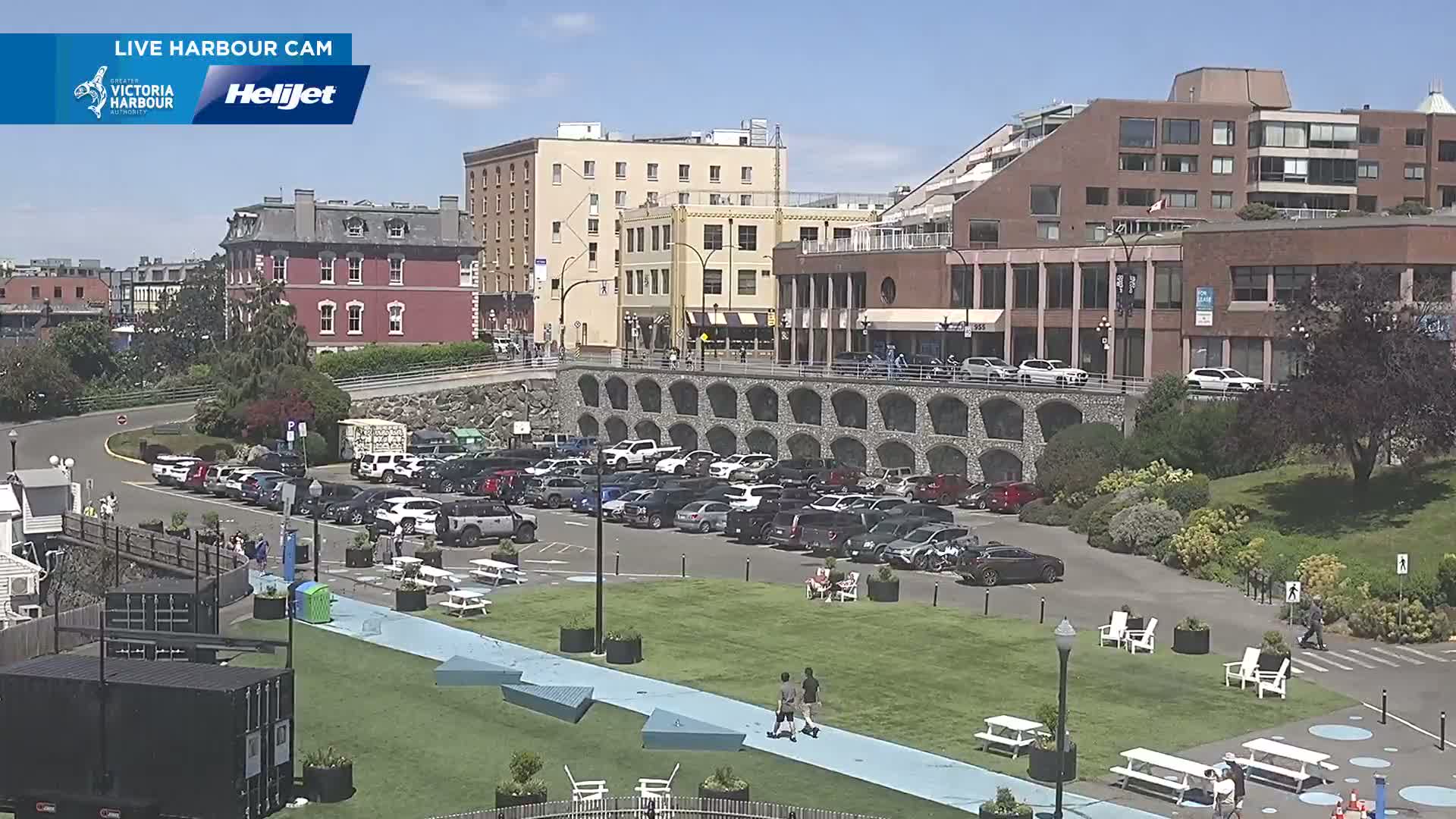 A sunny day reveals a waterfront scene with a large parking lot filled with cars, a grassy area with picnic tables and people strolling, and multi-story buildings in the background.