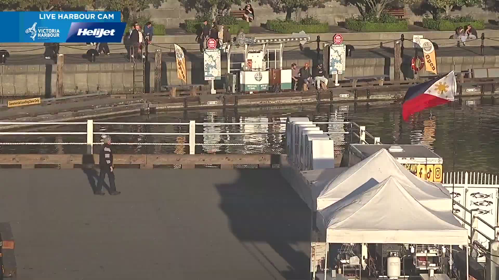 A waterfront scene on a sunny day shows a person walking along a dock near calm water, with several people standing near a small building, a few flags, and white canopy tents nearby.