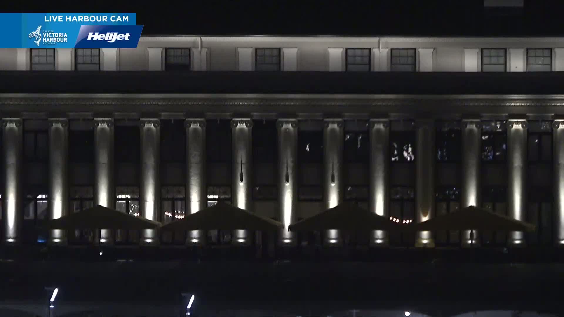 At night, a large, classically styled building with columns is illuminated, and several small, closed-top canopies sit in front of it.