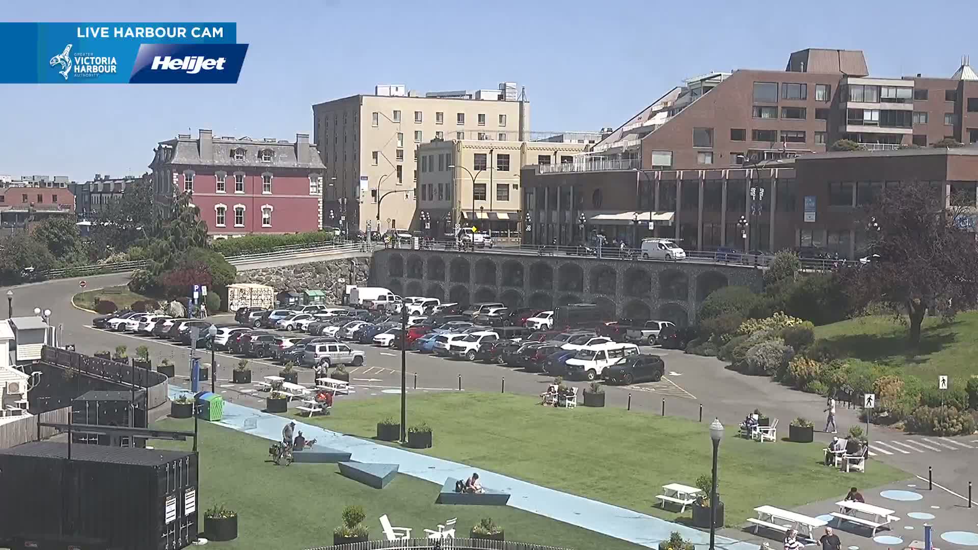 A sunny day overlooking a city harbor with a large parking lot, a grassy area with picnic tables and people relaxing, and several buildings in the background.