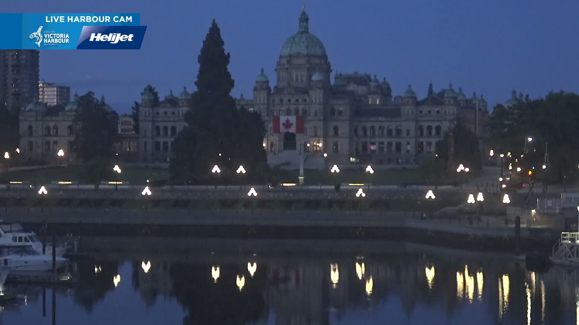 A large, stone building with a green dome is illuminated at night, reflecting in calm water, with boats docked nearby under a dark blue sky.