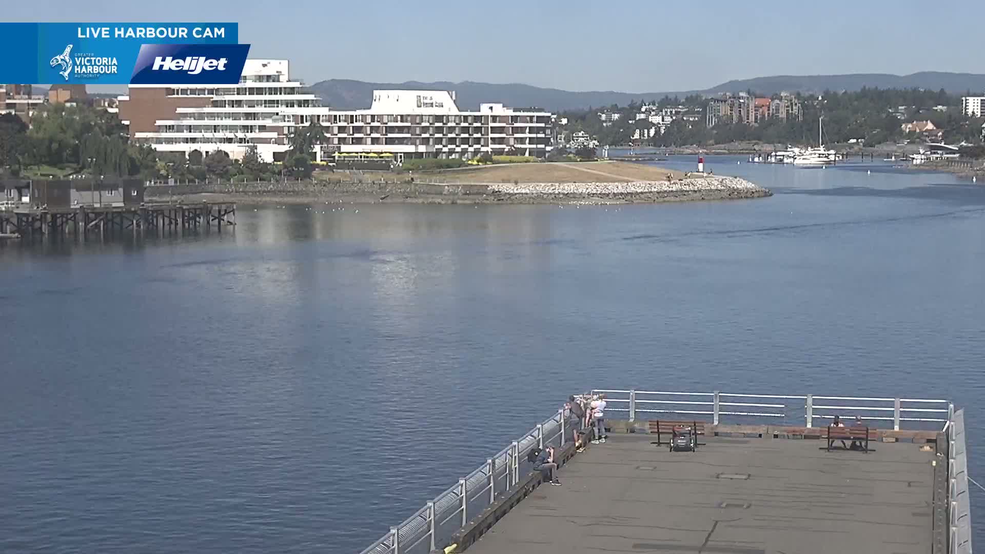 A sunny day shows a calm harbor with a large hotel in the background, viewed from a pier with several people sitting on benches.