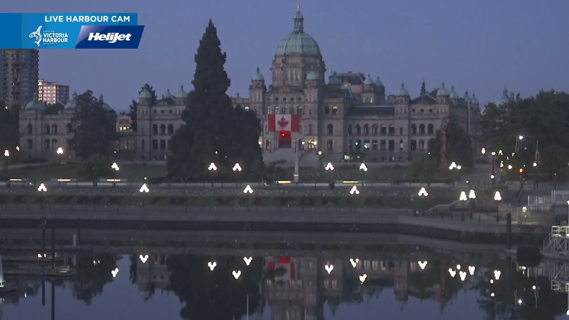 A large, ornate stone building with a green dome is illuminated at dusk, reflected in calm water, with lights lining a walkway in front.