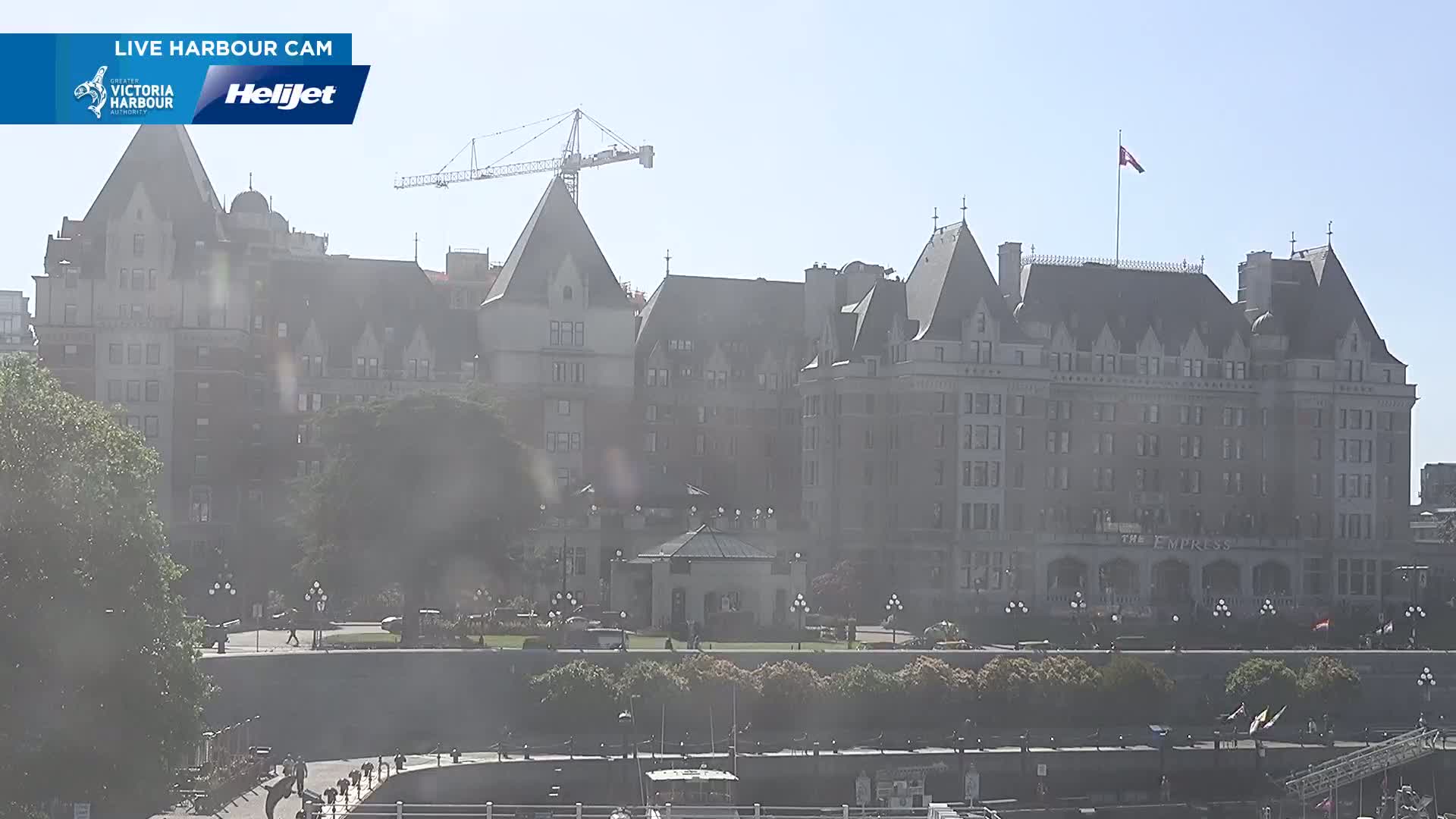 A large, historic hotel stands on a waterfront on a hazy, sunny day, with a construction crane visible in the background.