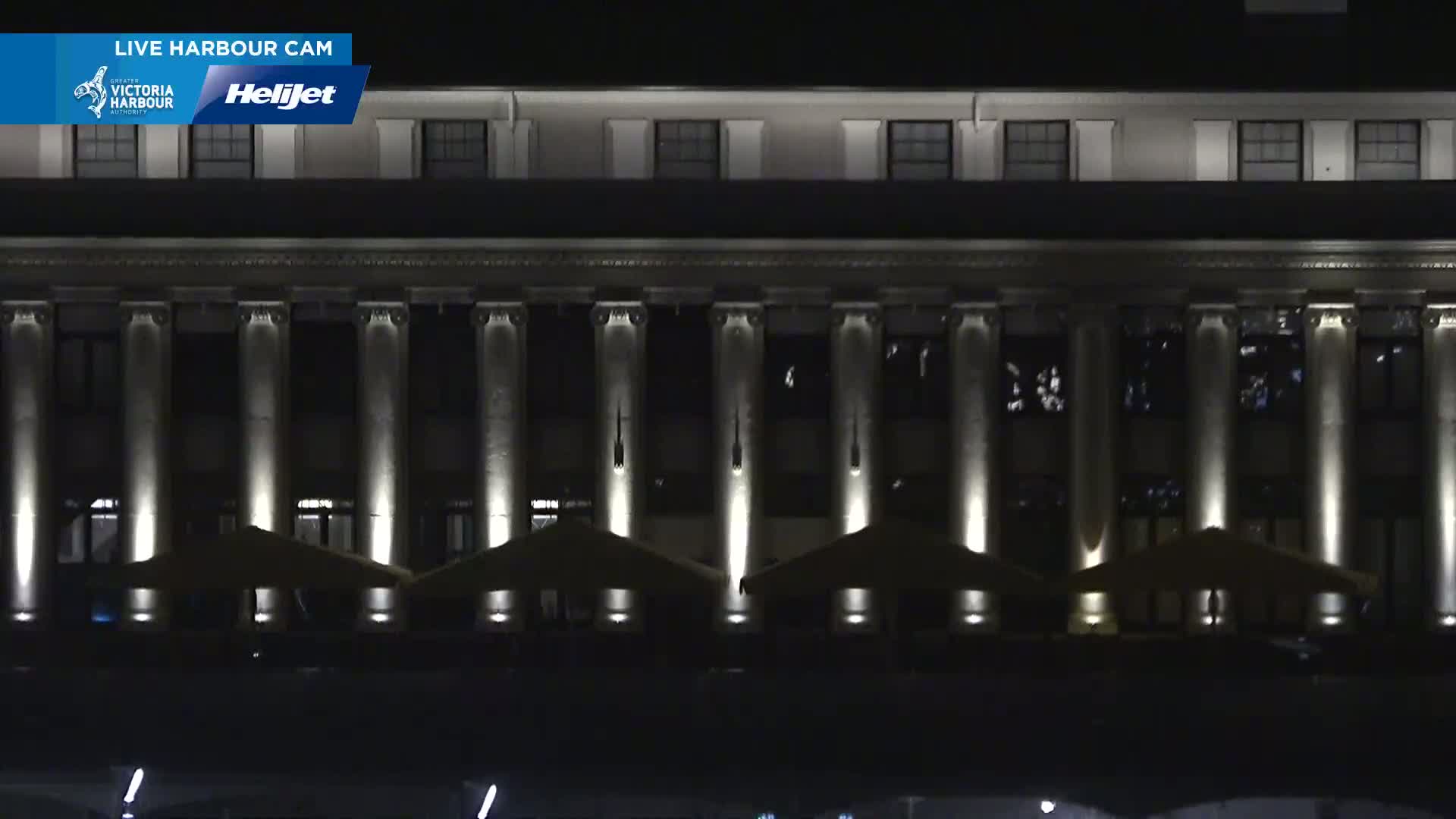 A large, classical-style building with many columns is illuminated at night, with several patio umbrellas visible in front.