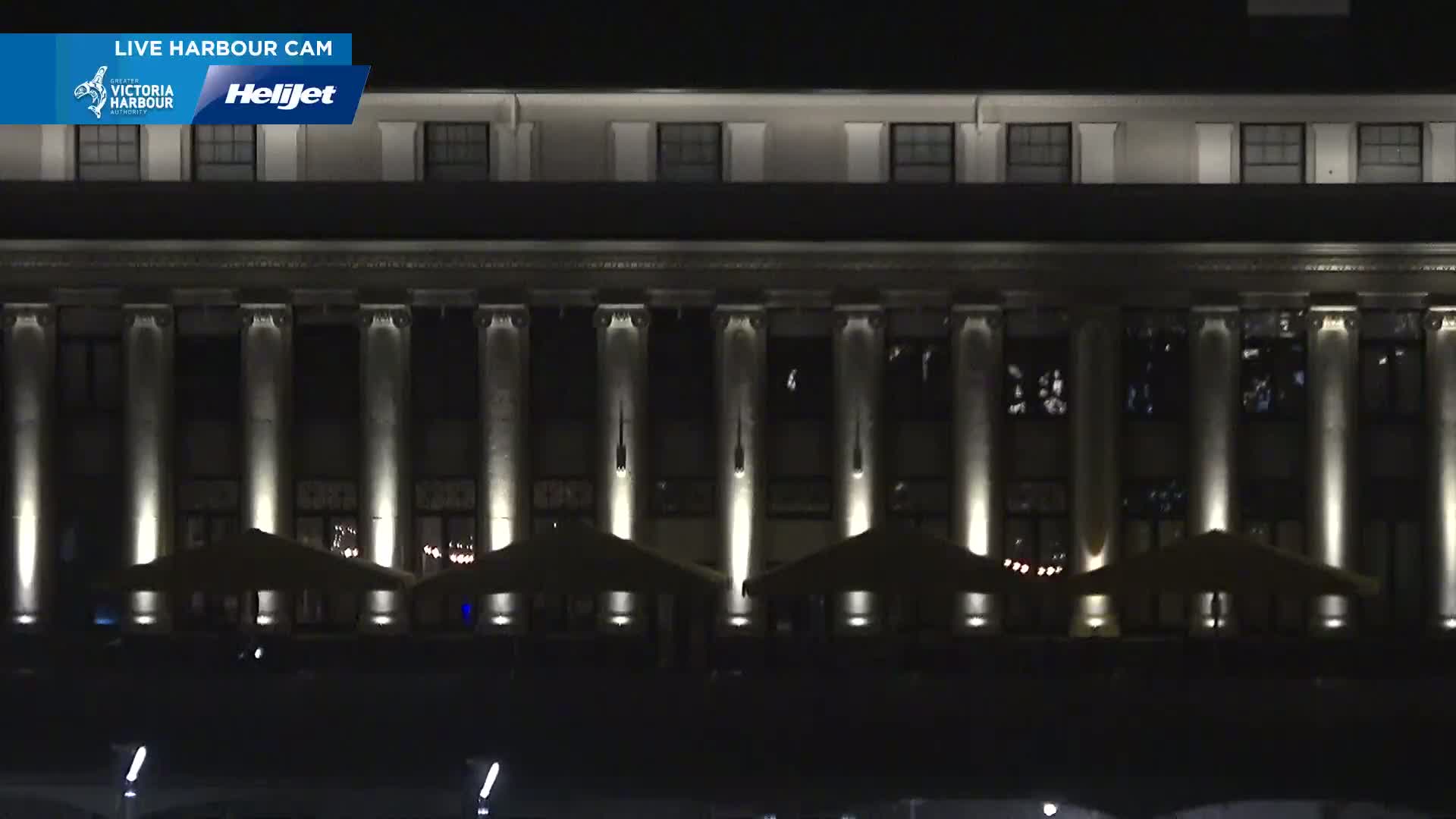 At night, a large, classical-style building with columns is illuminated, and several outdoor patio umbrellas are set up in front of it.