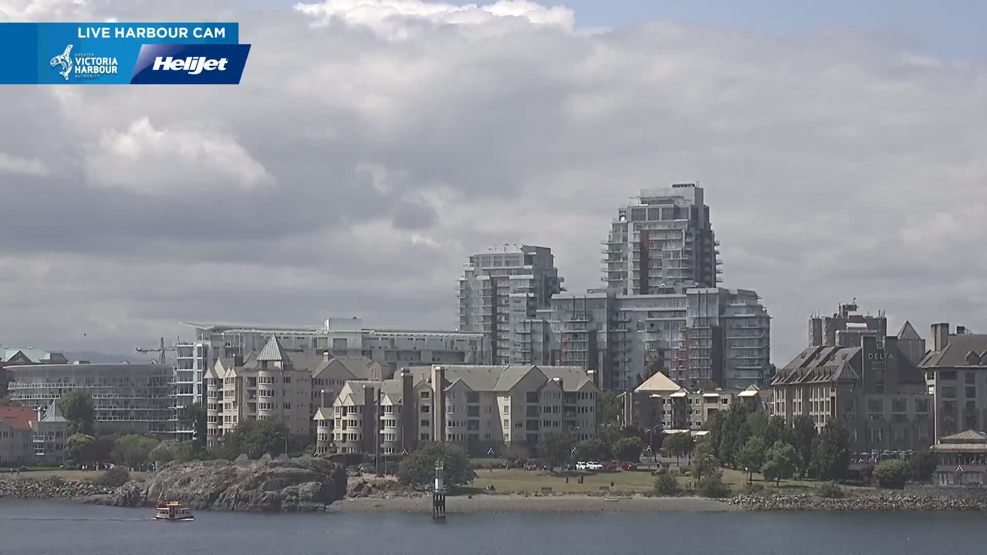 A waterfront city scene under a partly cloudy sky shows several multi-story buildings, some low-rise buildings, a small boat, and a rocky shoreline.