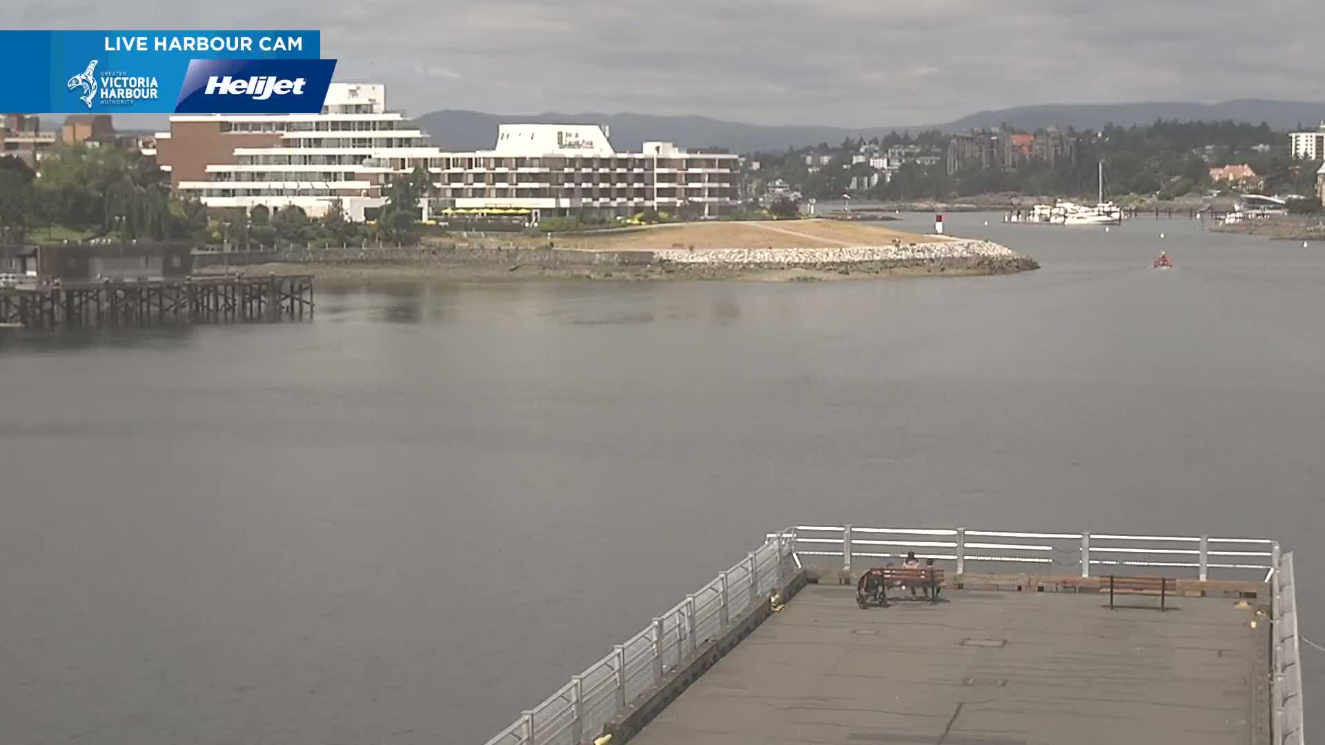 A calm, overcast day shows a harbor view with a large hotel, a small pier with two people sitting on a bench, and a larger pier in the background.