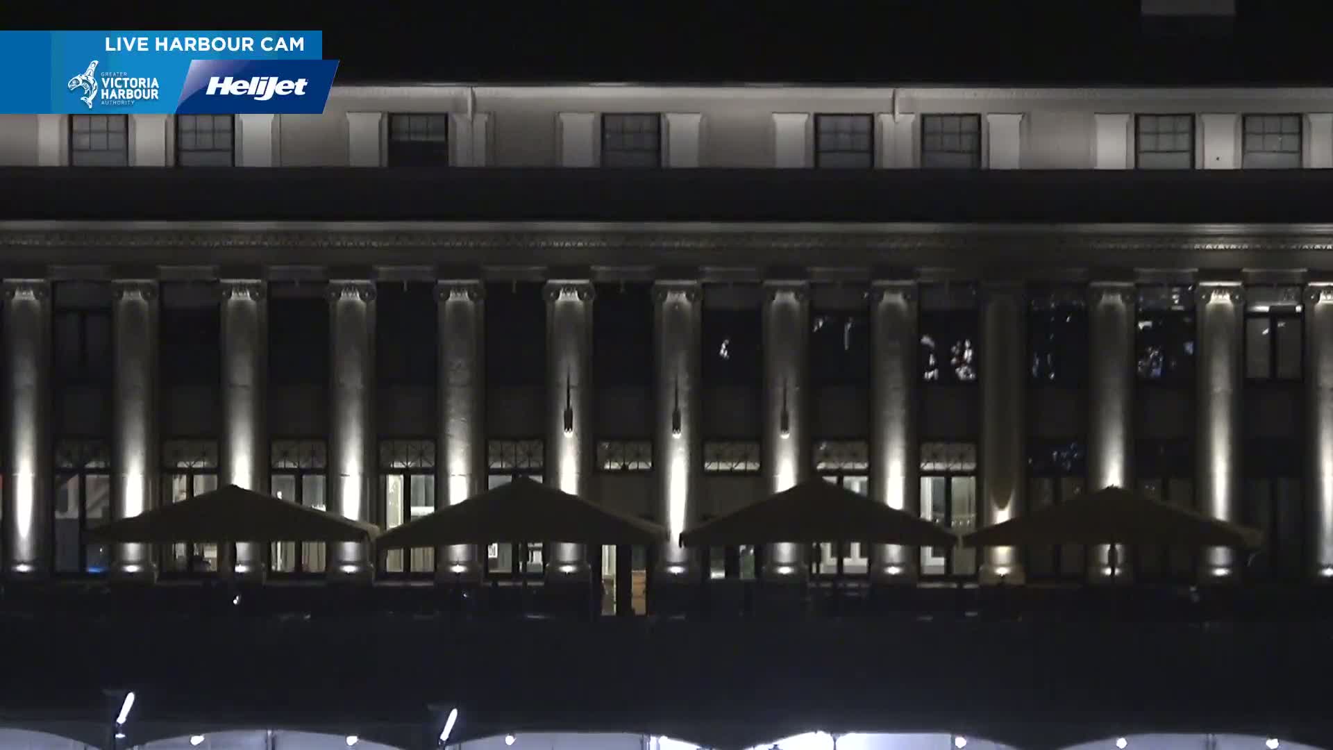A large, classical-style building with columns is illuminated at night, and several outdoor umbrellas are set up in front of it.