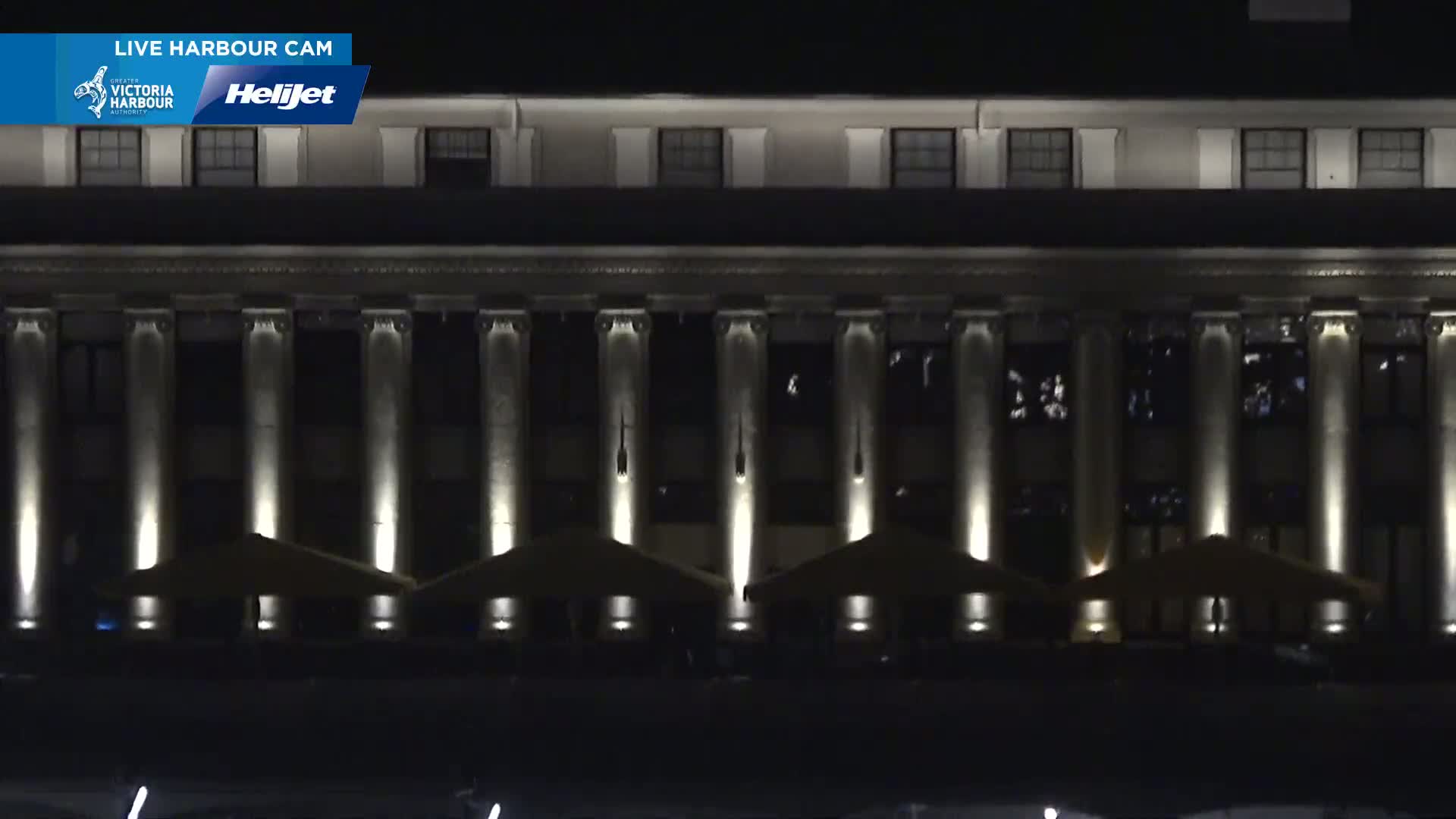 A large, classically styled building at night is illuminated, showing several columns and outdoor patio umbrellas.