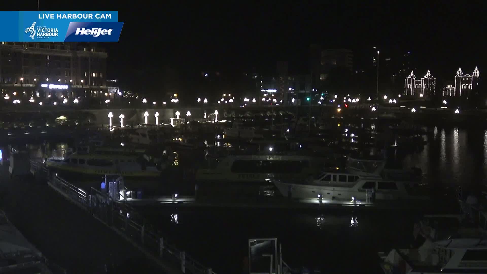 A nighttime view of a harbor filled with numerous boats moored alongside a waterfront, featuring illuminated buildings and a calm, dark body of water reflecting the lights.