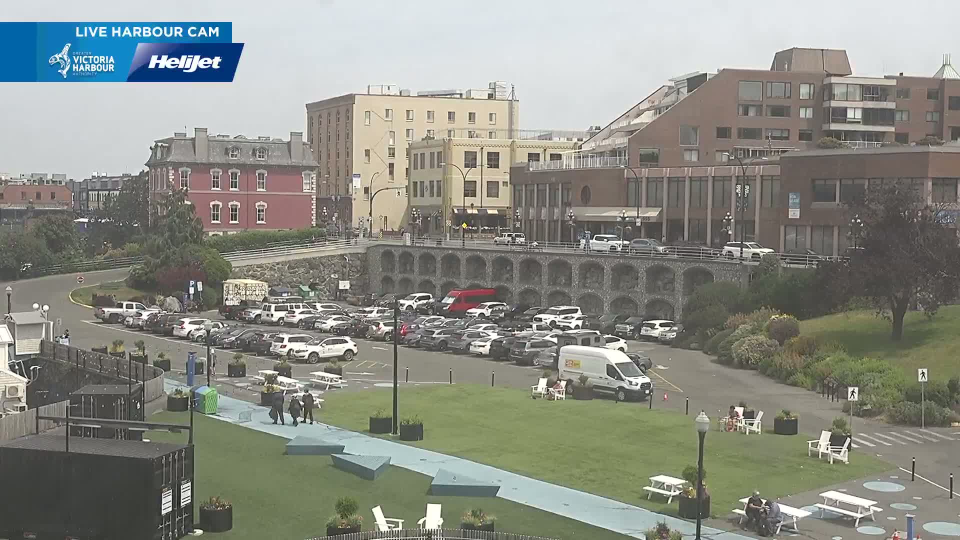A city harbor scene on a sunny day shows a parking lot full of cars, buildings, and a grassy area with picnic tables and people.