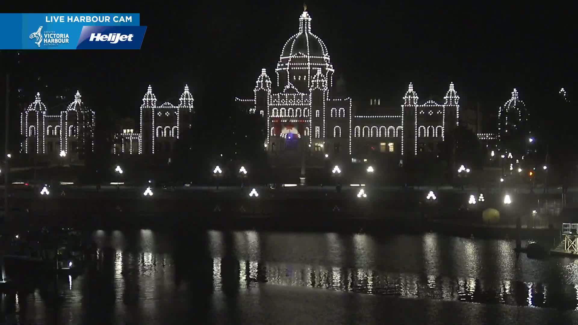 A large building, outlined in white lights, is reflected in dark water at night.