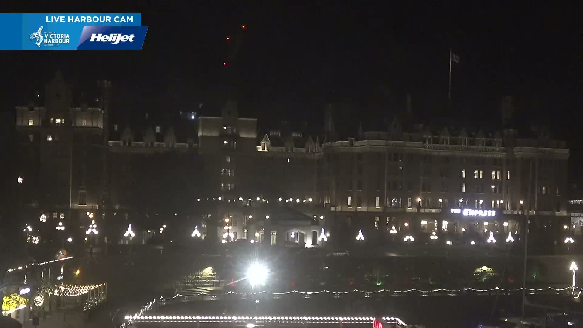 The image depicts a clear night scene of a well-lit waterfront featuring ornate historic buildings, including the prominently illuminated "The Empress" hotel, and numerous decorative lights strung along the foreground.