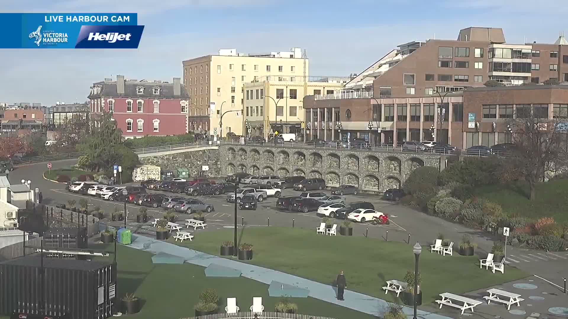A bright, sunny day illuminates a bustling city view featuring a green park space with picnic tables and a lone walker, a large parking lot filled with cars, and various multi-story buildings, including a prominent red brick structure, all beneath a mostly clear blue sky.