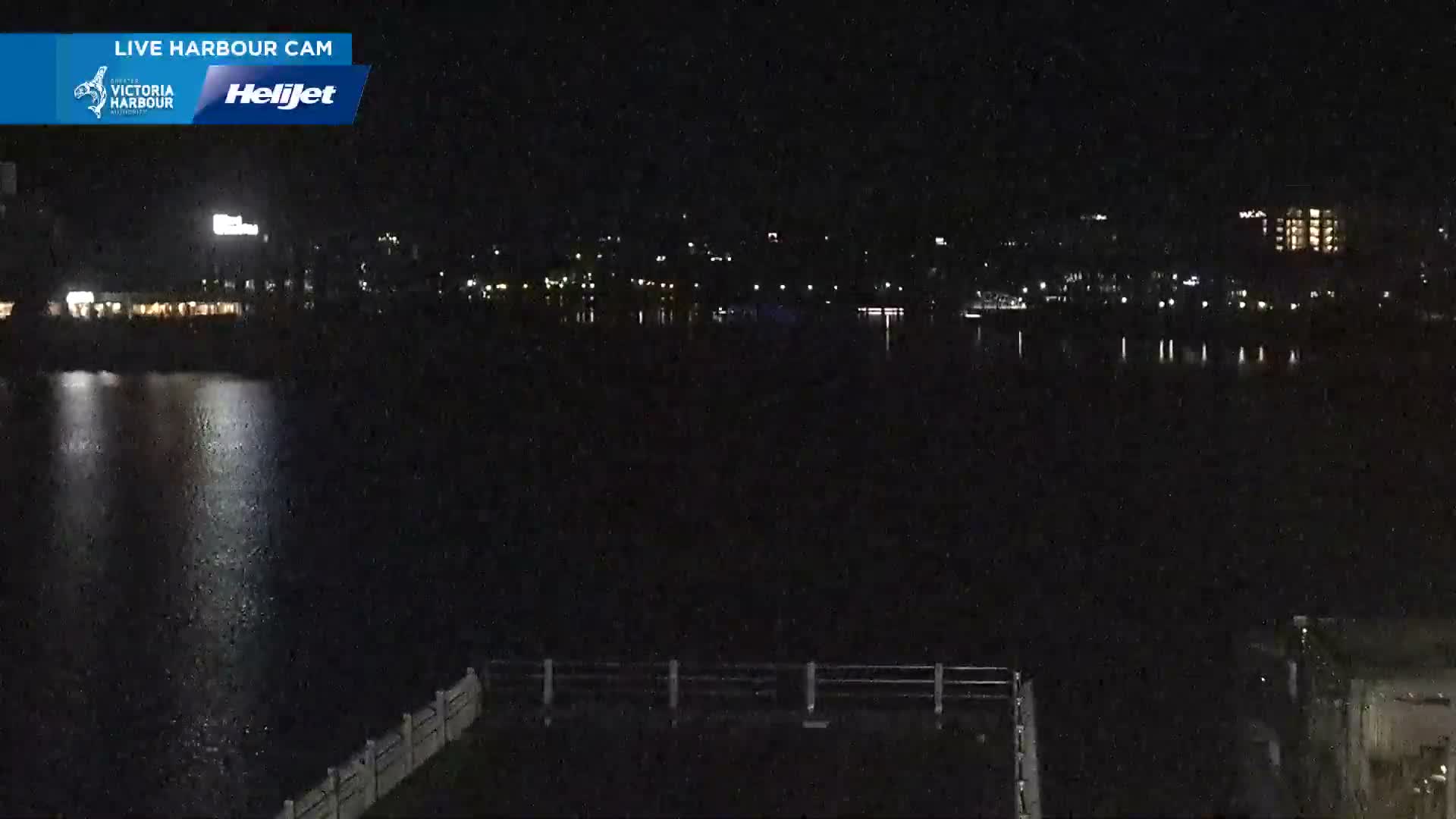 A bright, sunny day illuminates a bustling city view featuring a green park space with picnic tables and a lone walker, a large parking lot filled with cars, and various multi-story buildings, including a prominent red brick structure, all beneath a mostly clear blue sky.