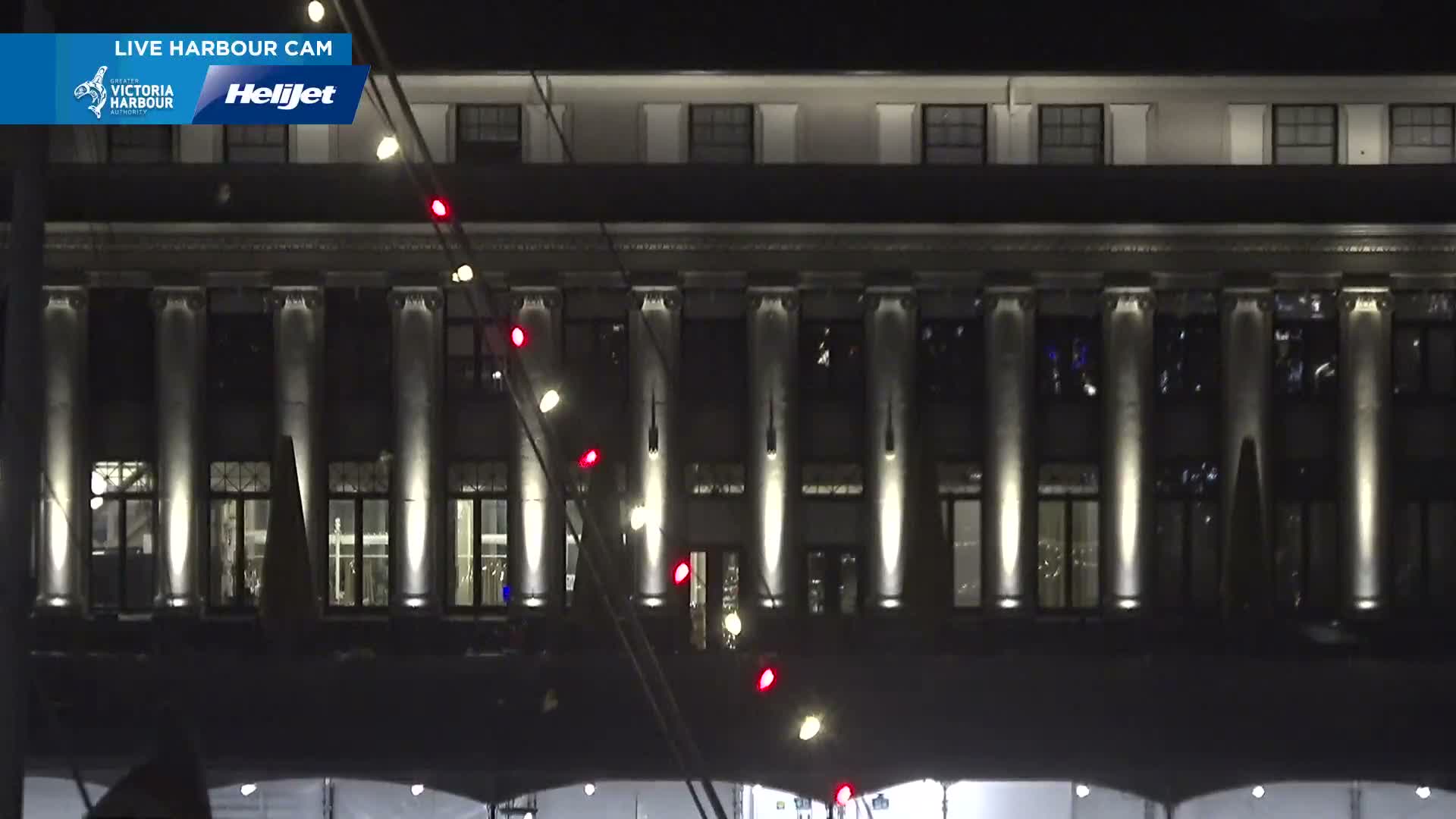 A grand building with illuminated columns and windows is seen at night under clear skies, with a diagonal string of festive red and white lights in the foreground.