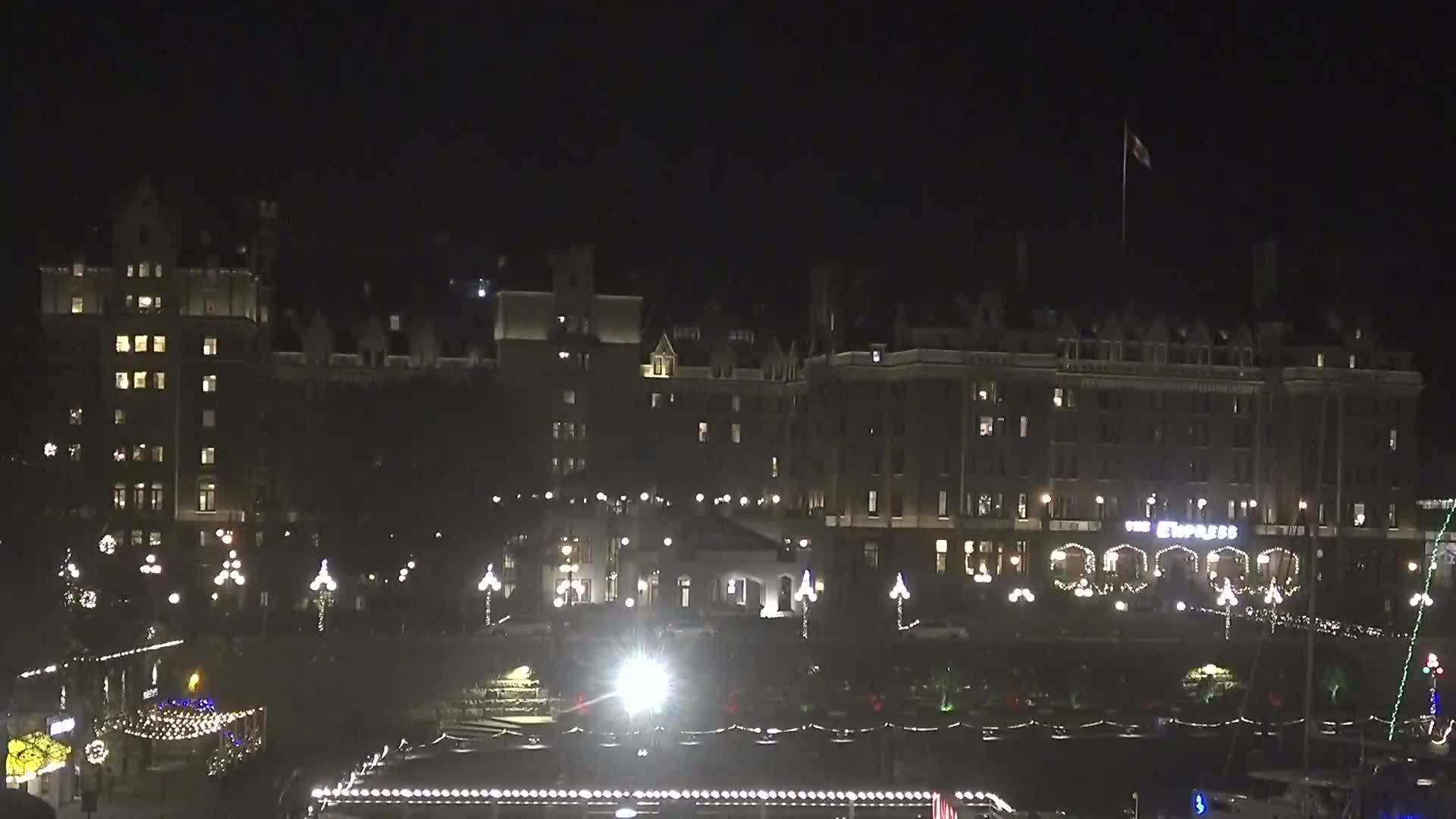 The image displays a large, ornate building illuminated at night with many lights, overlooking a festive waterfront area adorned with numerous bright decorations, all under a clear sky.