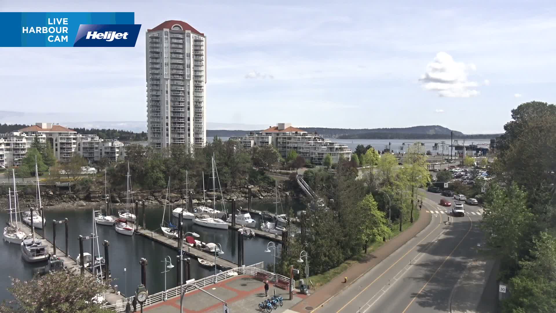 A marina filled with sailboats is situated beside a road and residential buildings, all under a partly cloudy sky.