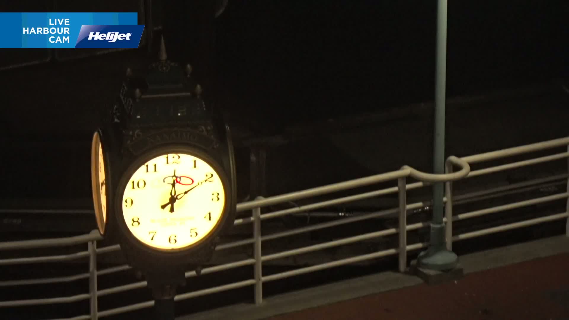 An illuminated clock hangs on a post beside a railing at night.