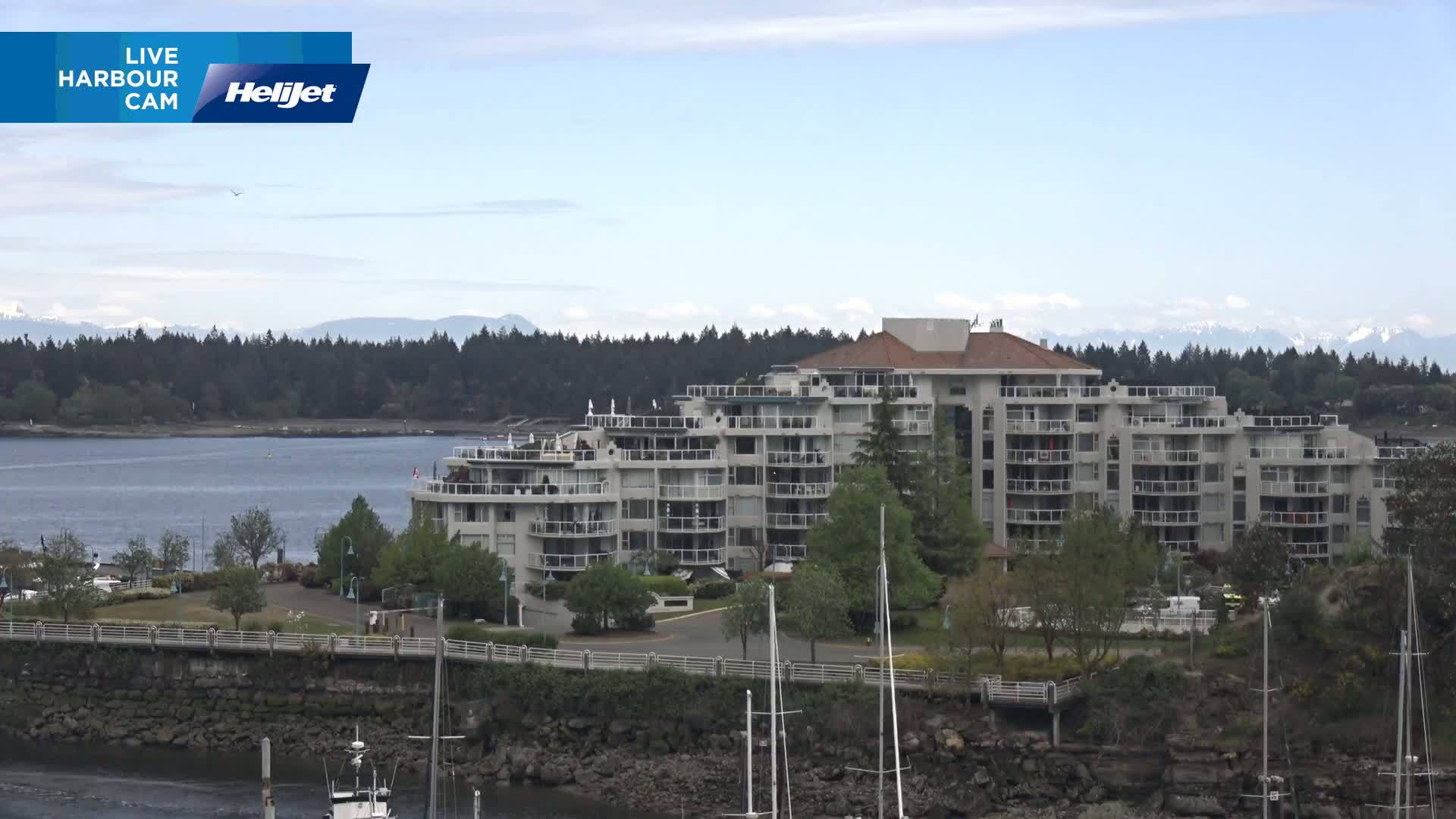A multi-story apartment building sits on a waterfront, surrounded by trees and a calm body of water with mountains in the background under a partly cloudy sky.
