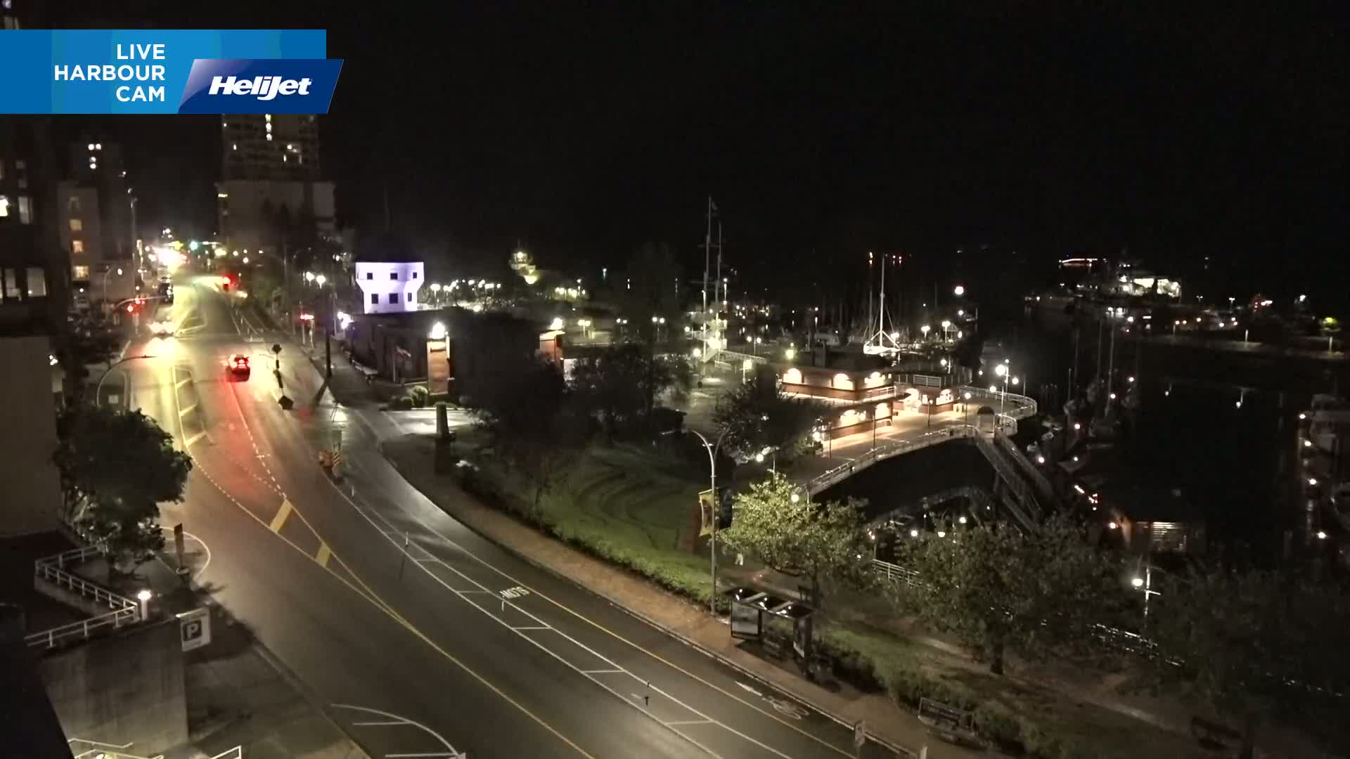 A nighttime, high-angle view shows a harbor with boats docked, a roadway with minimal traffic, and surrounding buildings and greenery.