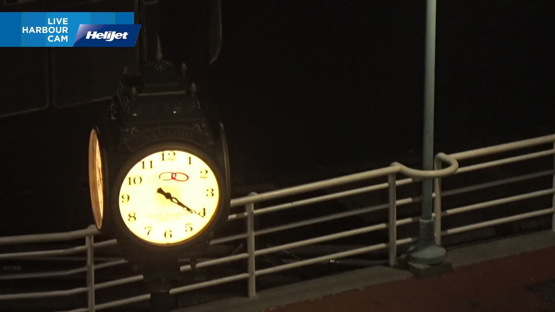 At night, a lighted clock hangs from a post in front of a railing.