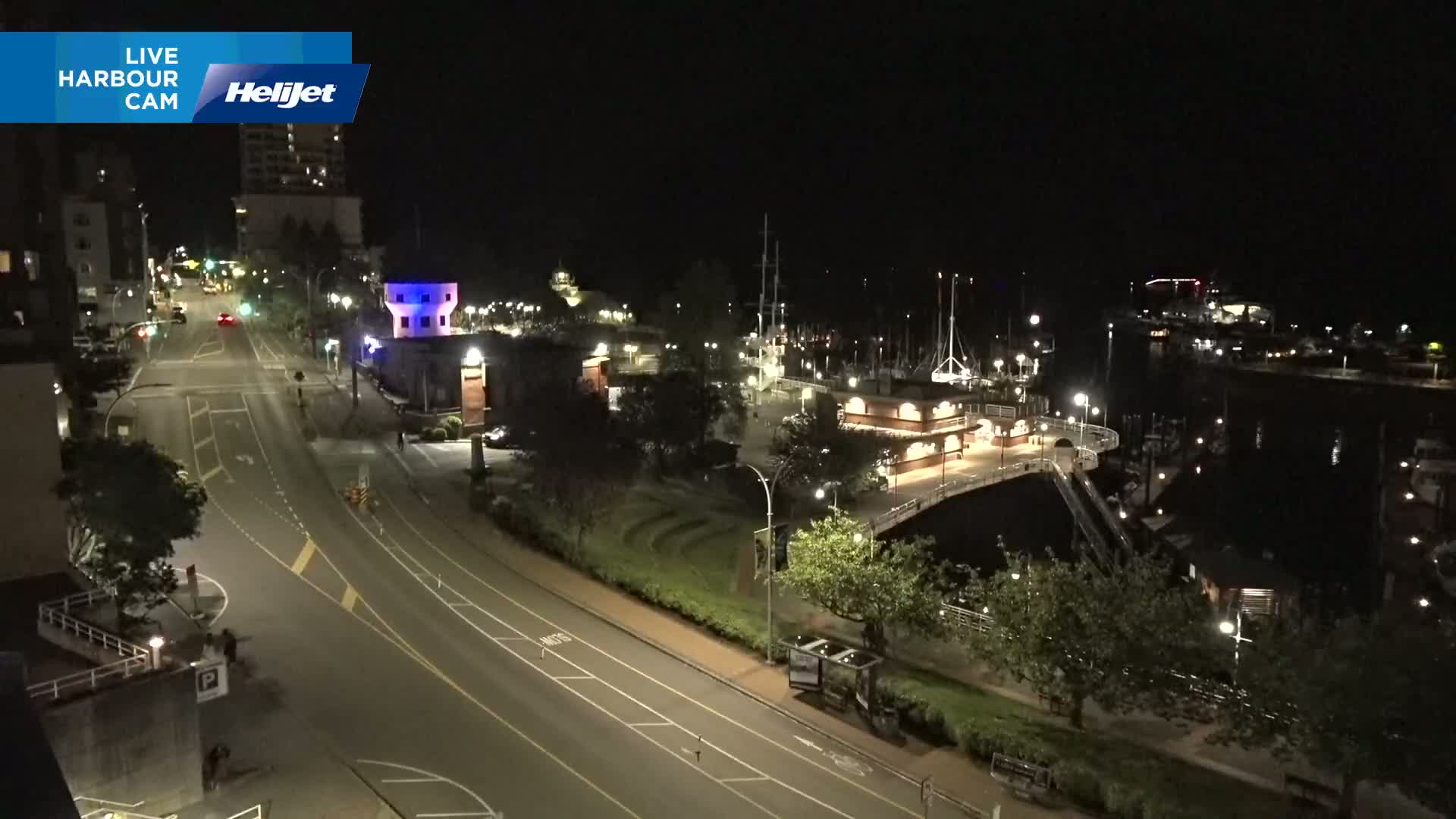 A nighttime, aerial view of a harbor with docked boats, a roadway, and buildings under clear skies.