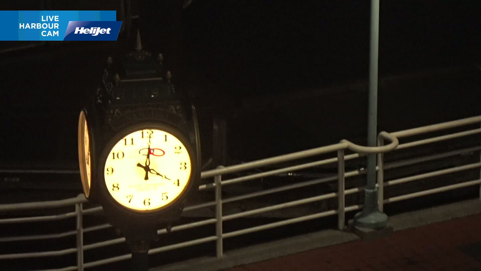 A brightly lit clock on a dark night is mounted on a post beside a railing.
