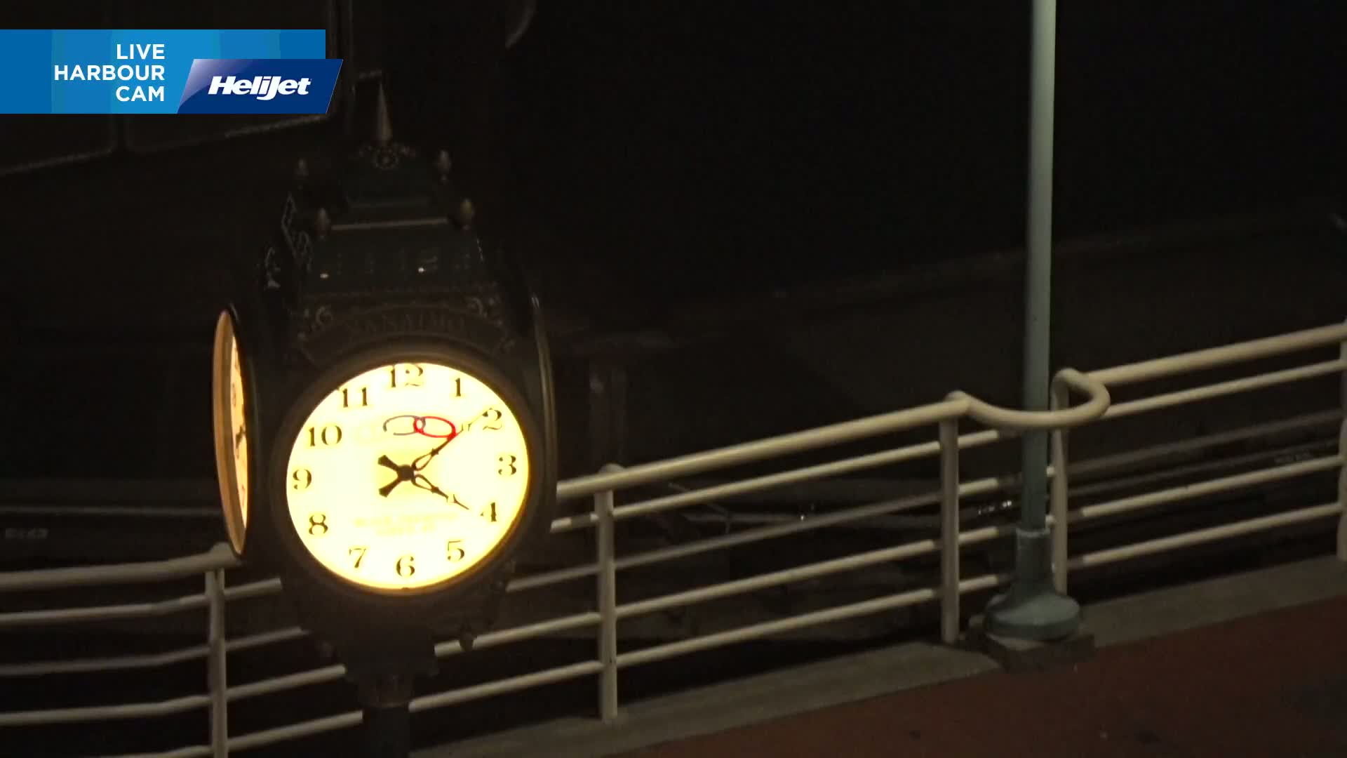 A brightly lit clock on a dark night is mounted on a post behind a white railing.