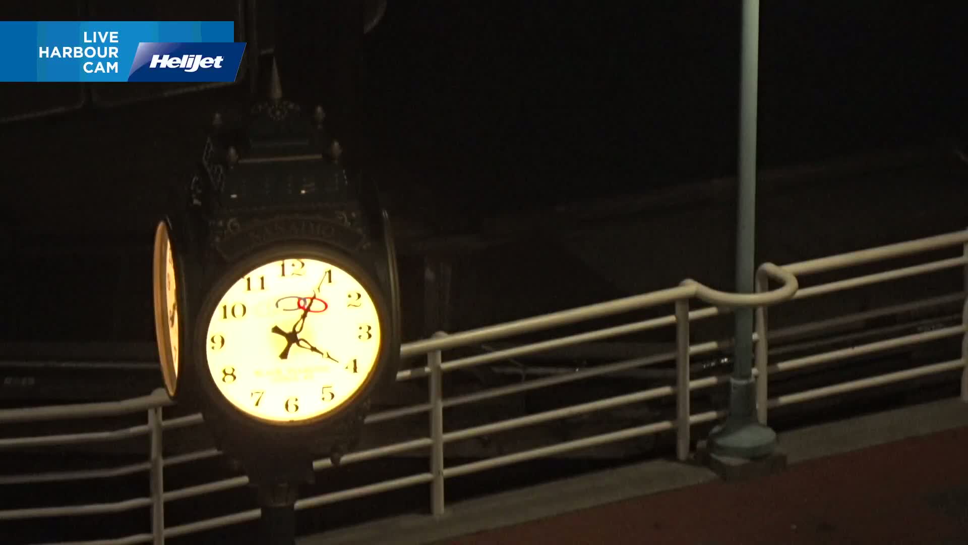 A brightly lit, old-fashioned clock is mounted on a post next to a railing at night.