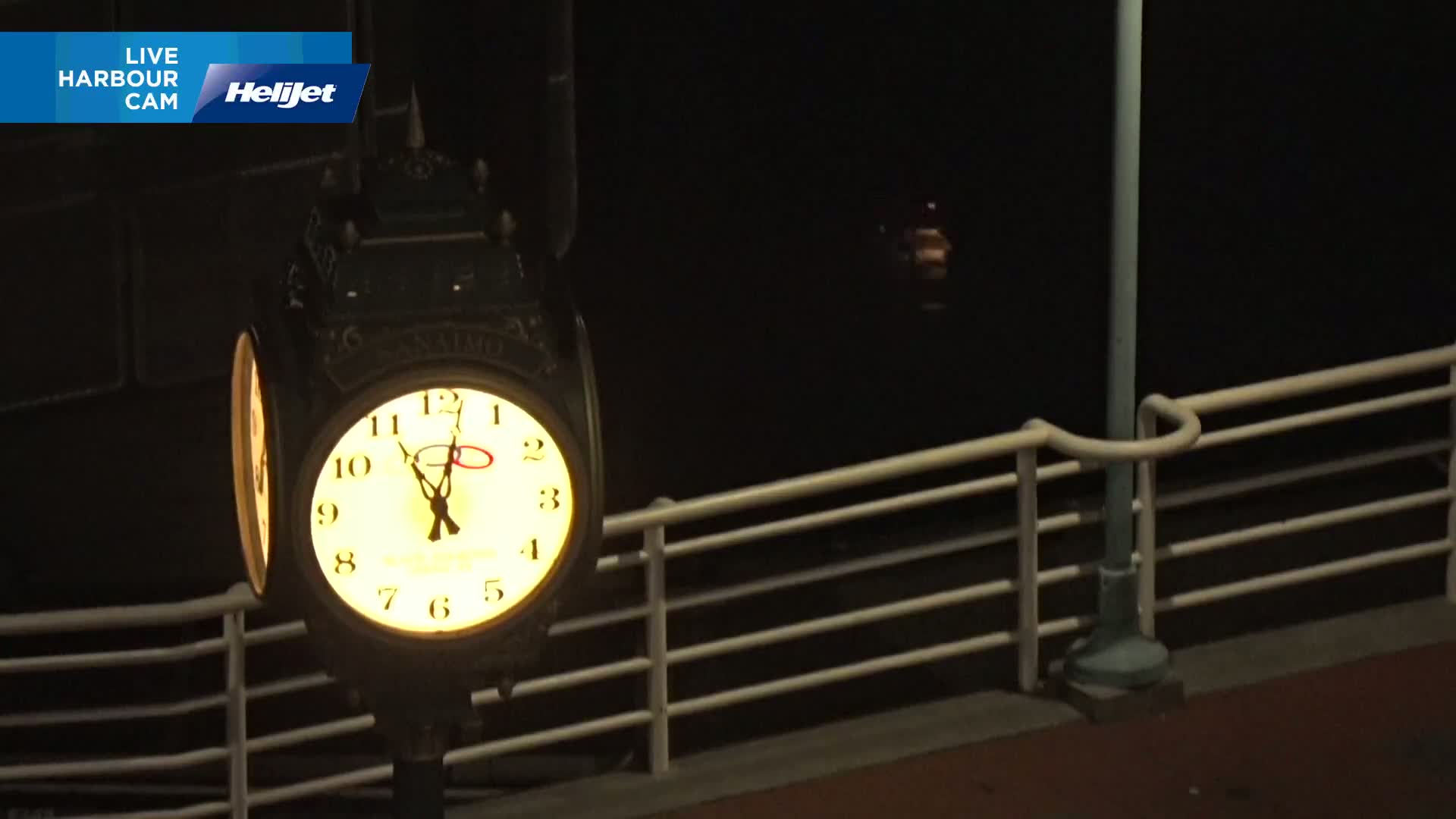 At night, an illuminated clock hangs on a post next to a railing overlooking dark water.