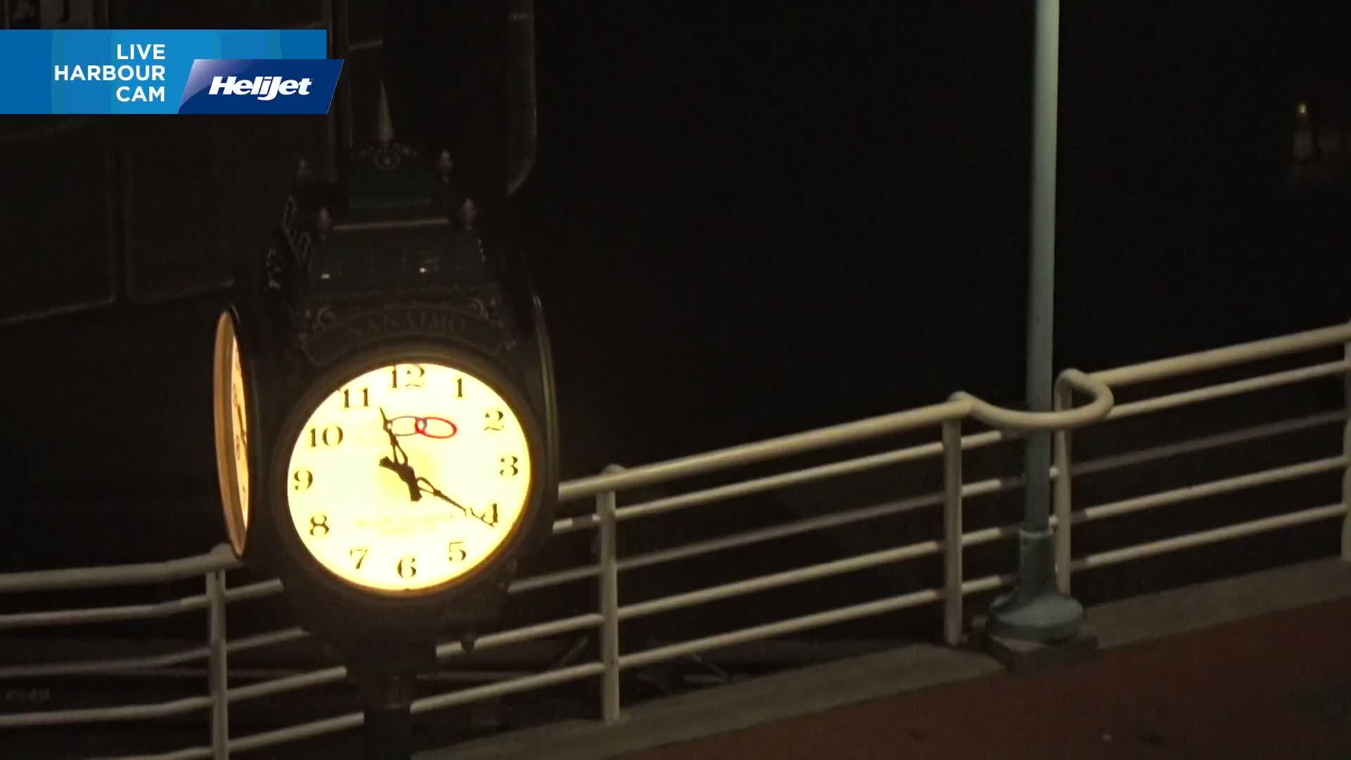 A lit, antique-style clock is mounted on a post beside a railing at night.